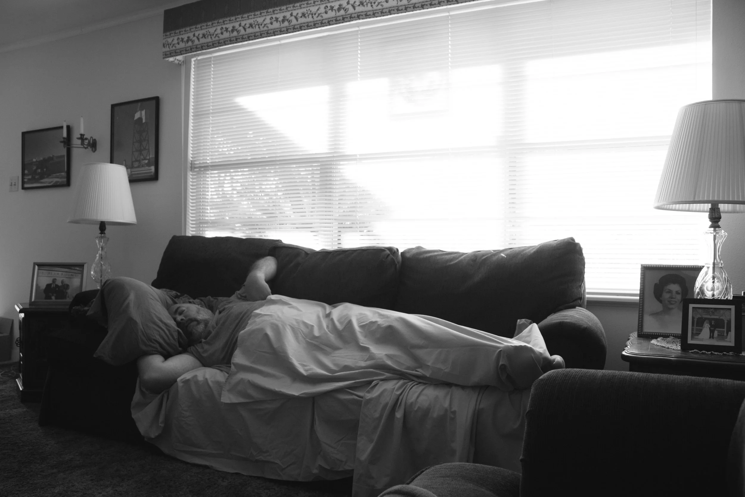 A man sleeping on a couch in a living room, with framed photographs on side tables, and window blinds letting in natural light.