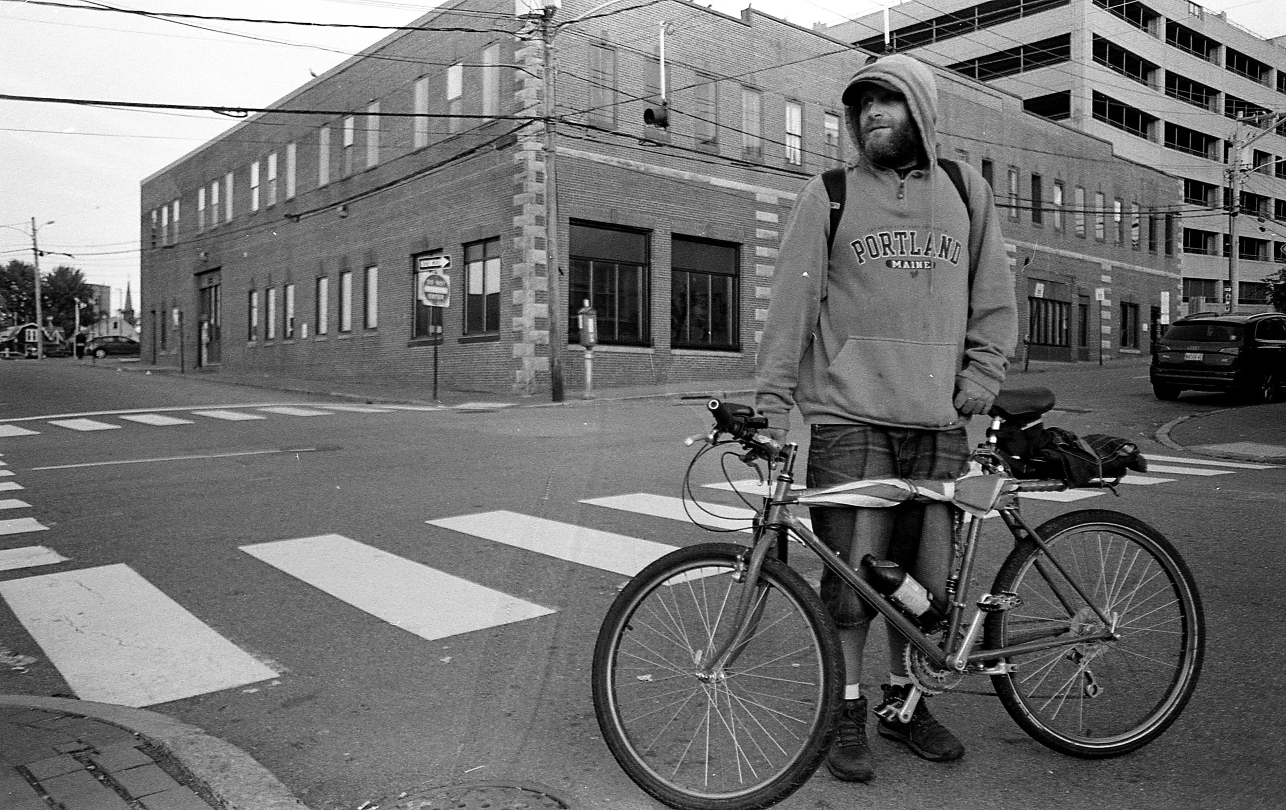 A man standing next to a bicycle on a city street corner in black and white, with a brick building, power lines, and a parking lot in the background.