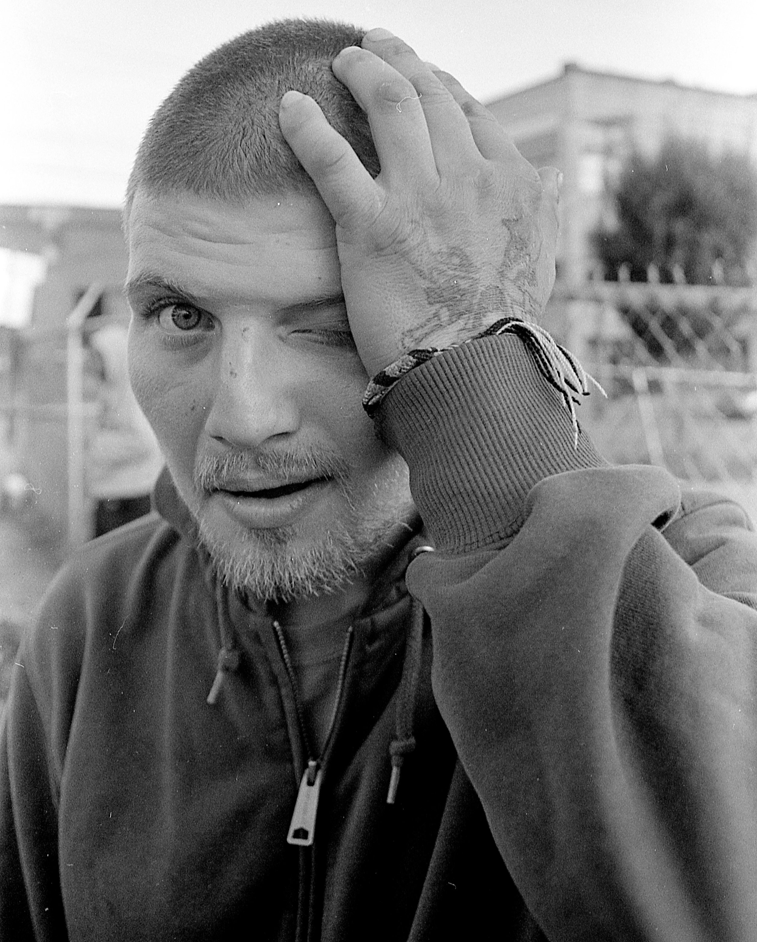 A man with a beard and short hair holding his head with his left hand, one eye closed, in a close-up black and white photo outdoors.