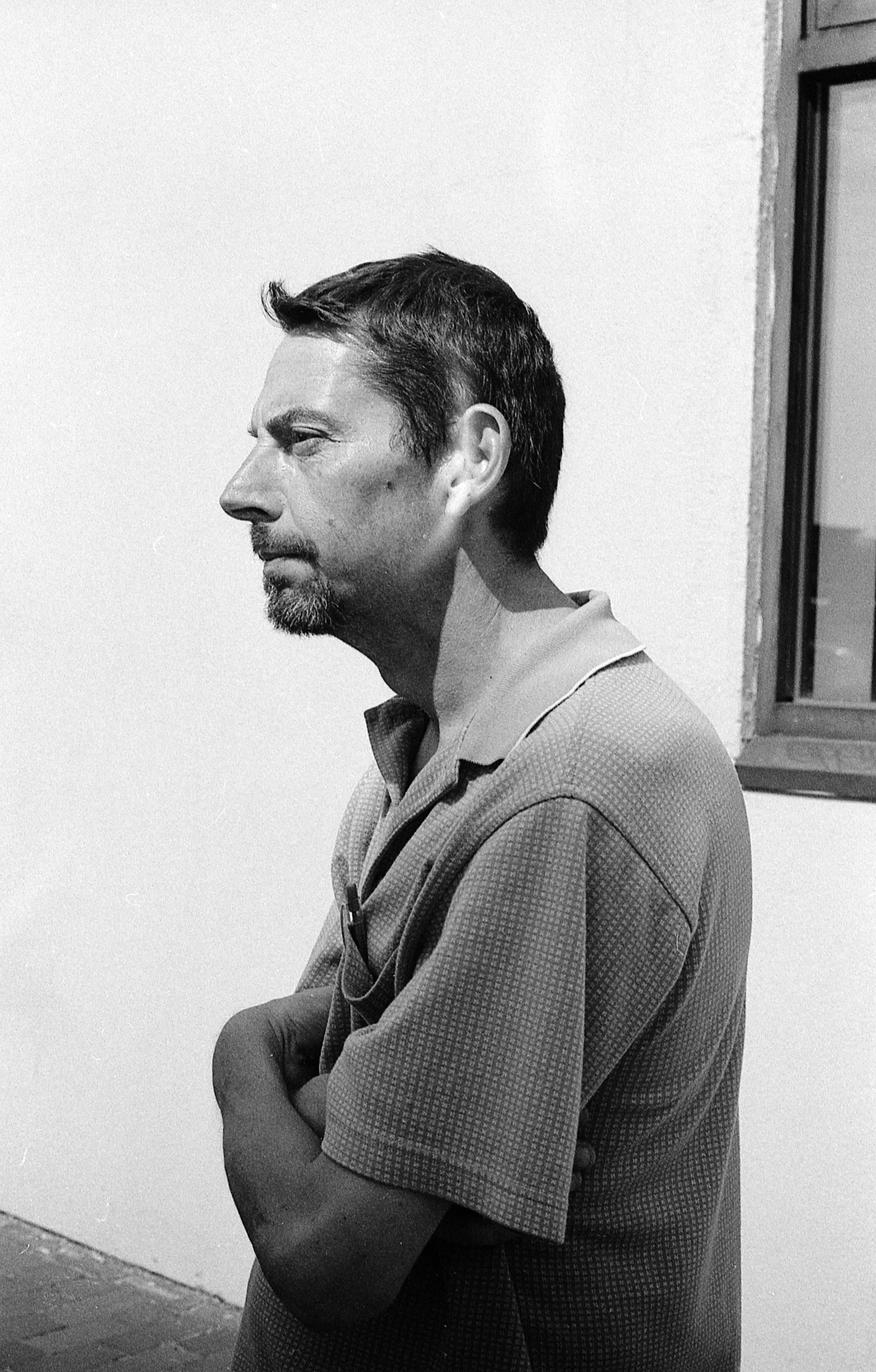 A black-and-white photo of a man in profile, standing against a plain wall with a window nearby, wearing a collared shirt and showing a serious expression.