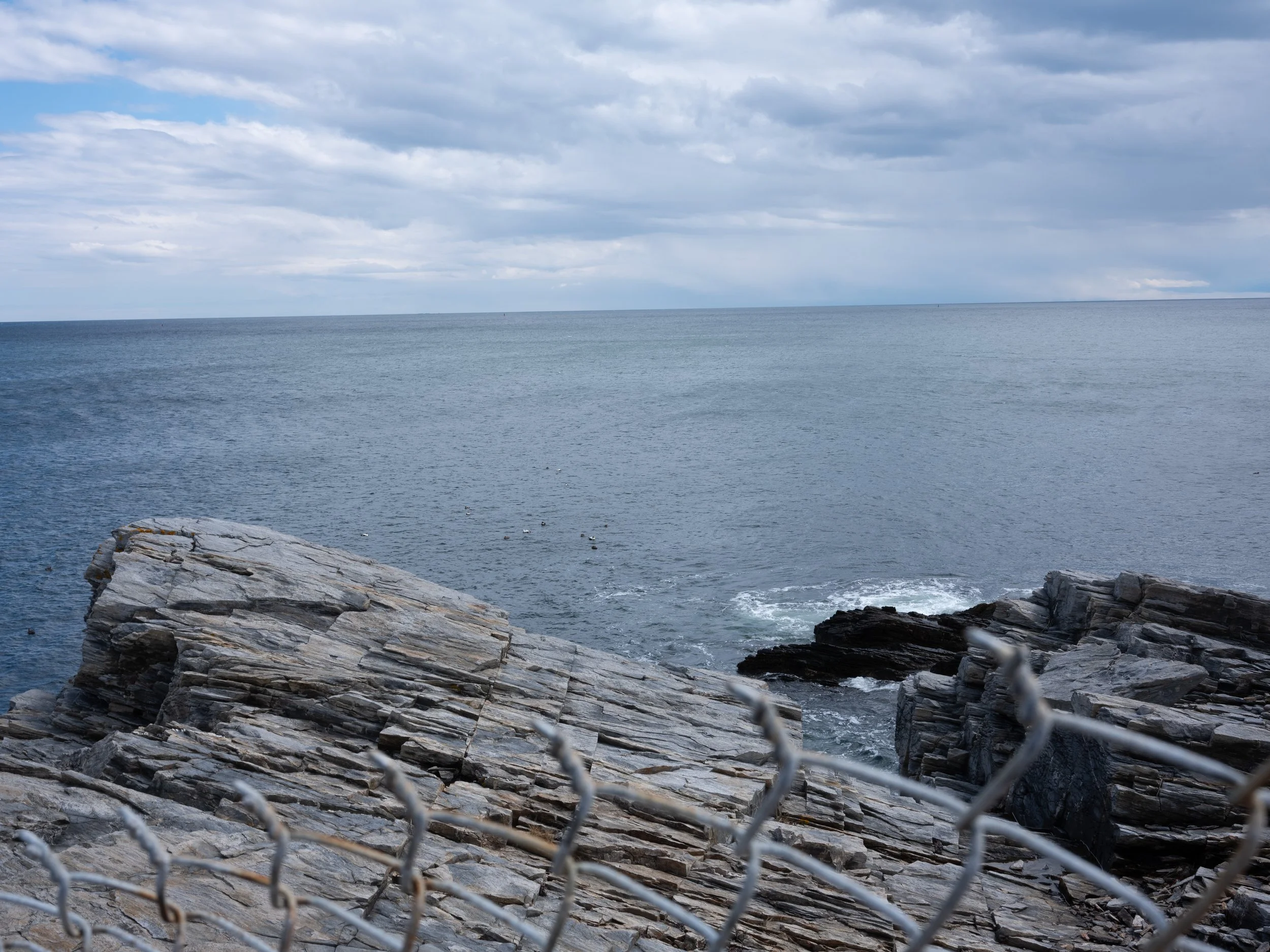 View of the ocean with rocky shoreline and cloudy sky, with a chain-link fence in the foreground.