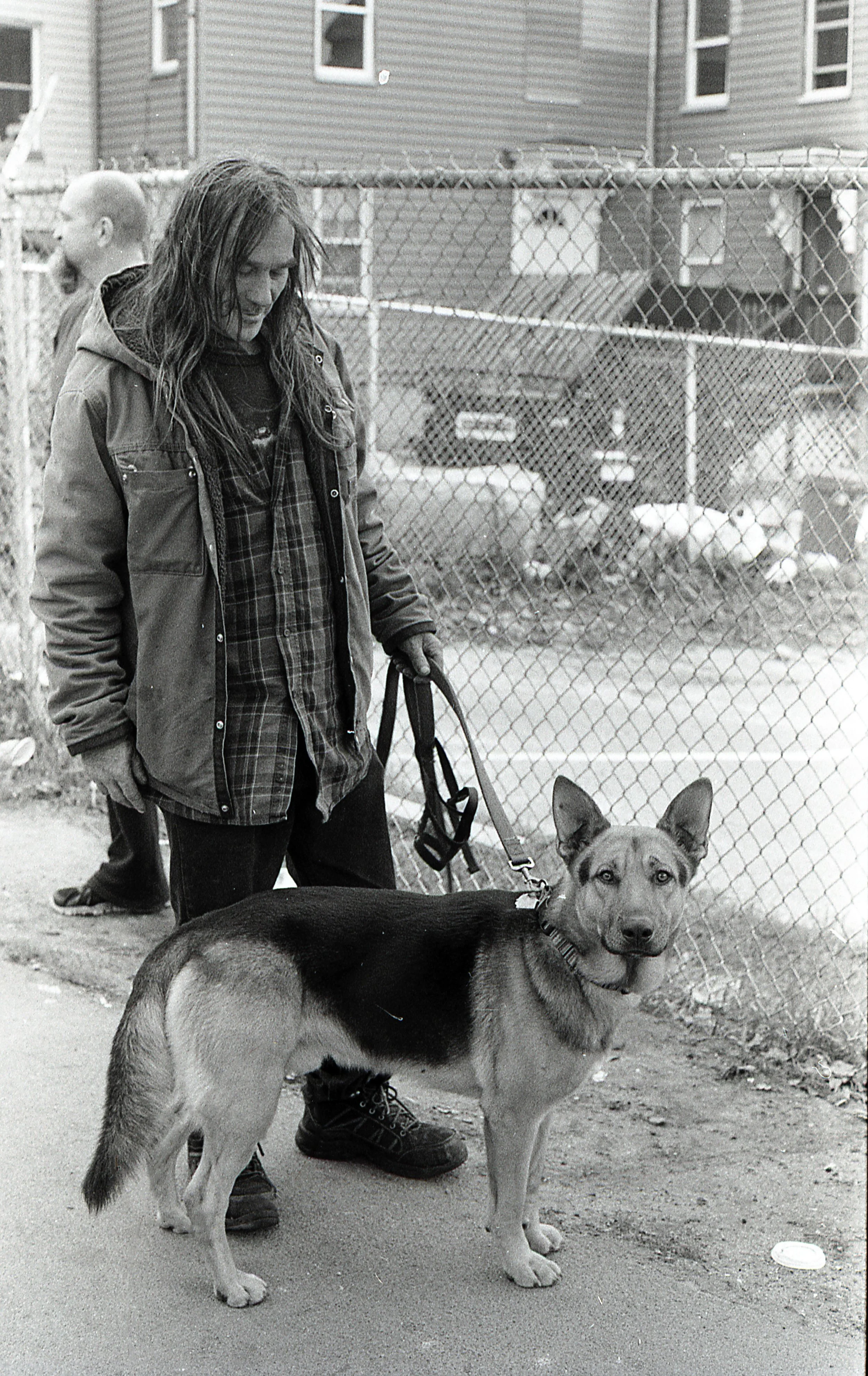 A woman with long hair wearing a jacket and gloves holds a dog on a leash. The dog appears to be a young mixed breed with erect ears and a black and tan coat, standing on a sidewalk in front of a chain-link fence.
