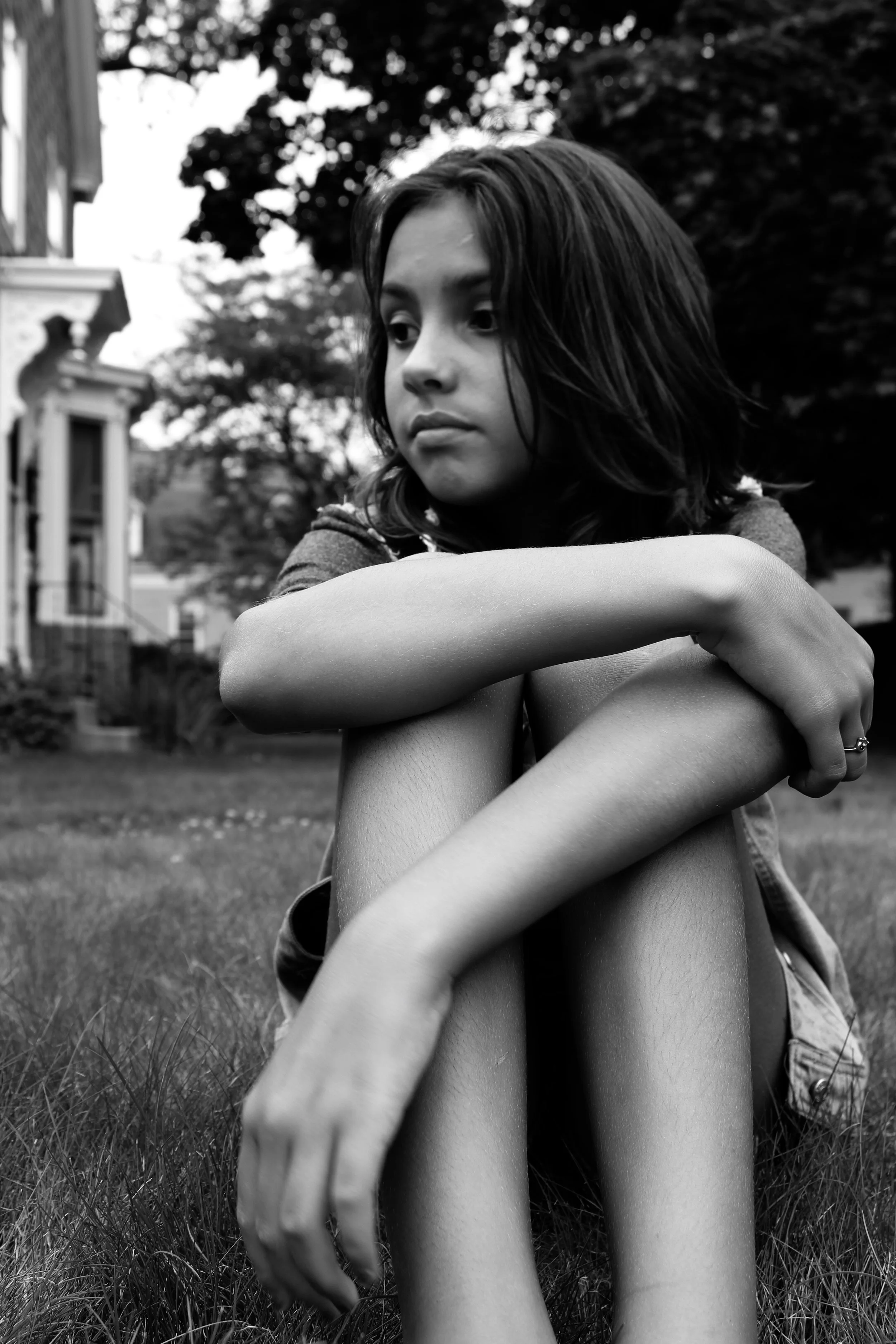 A young girl with shoulder-length hair sitting on grass, looking thoughtfully into the distance with her arms wrapped around her knees, in front of a house and trees.