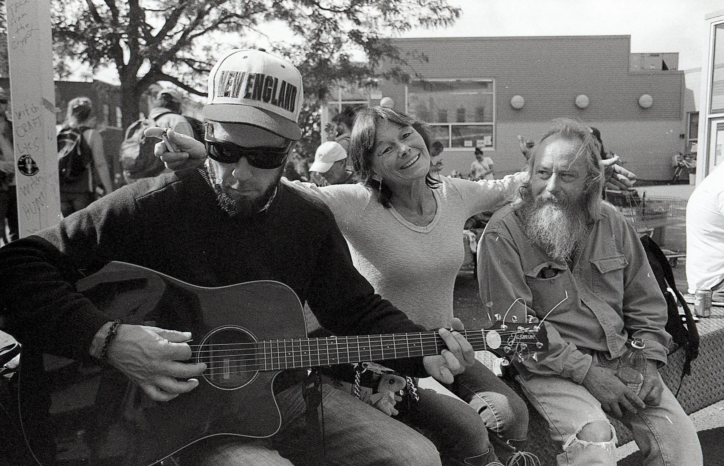 Three people sitting outdoors, one playing an acoustic guitar, a woman smiling with her arms around two men, one of whom has a long beard and long hair, the other wearing sunglasses and a cap that says "New England."