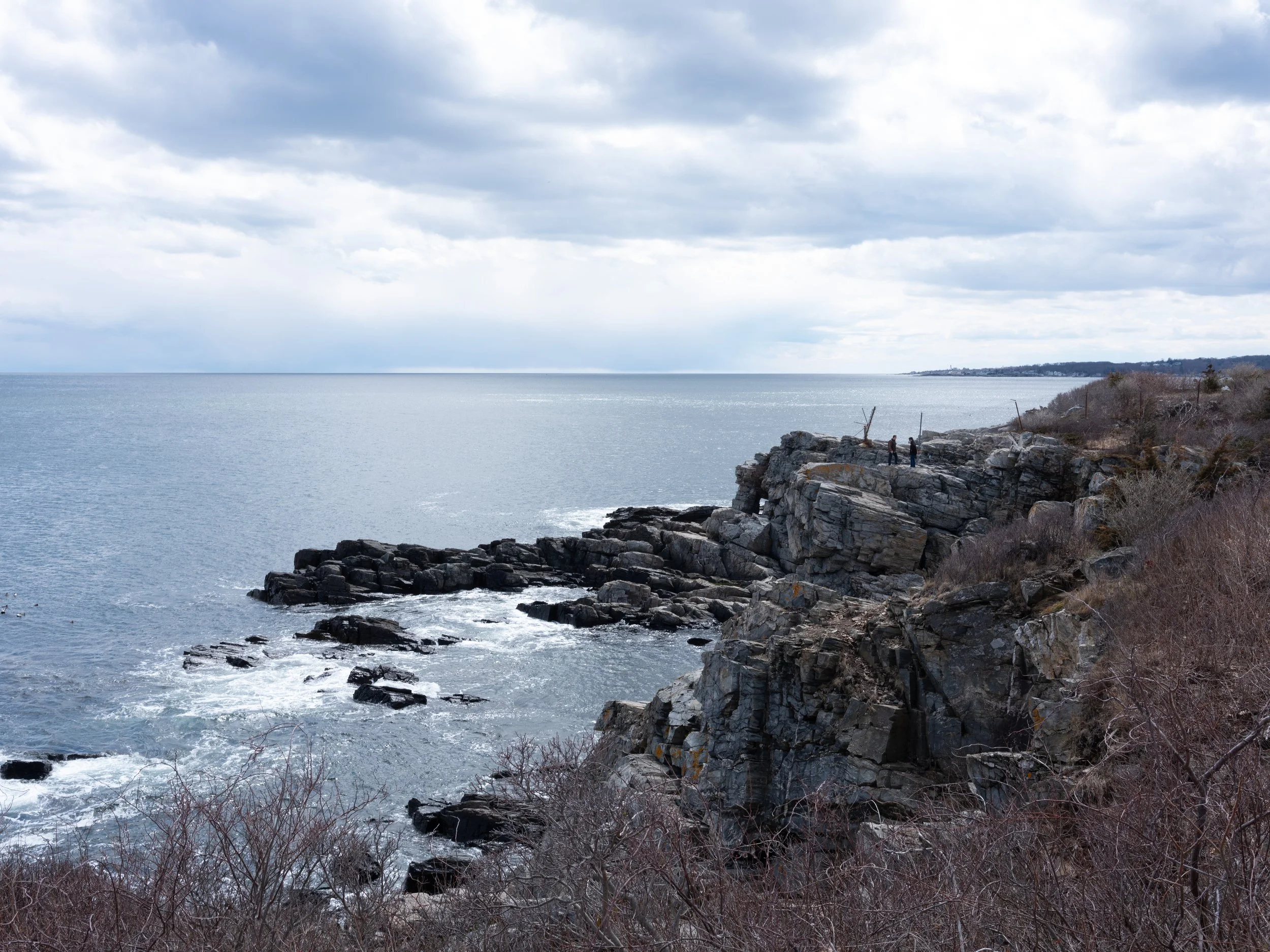 A rugged coastline with rocky cliffs and sparse vegetation, overlooking the ocean under a cloudy sky, with a few people standing on the rocks in the distance.
