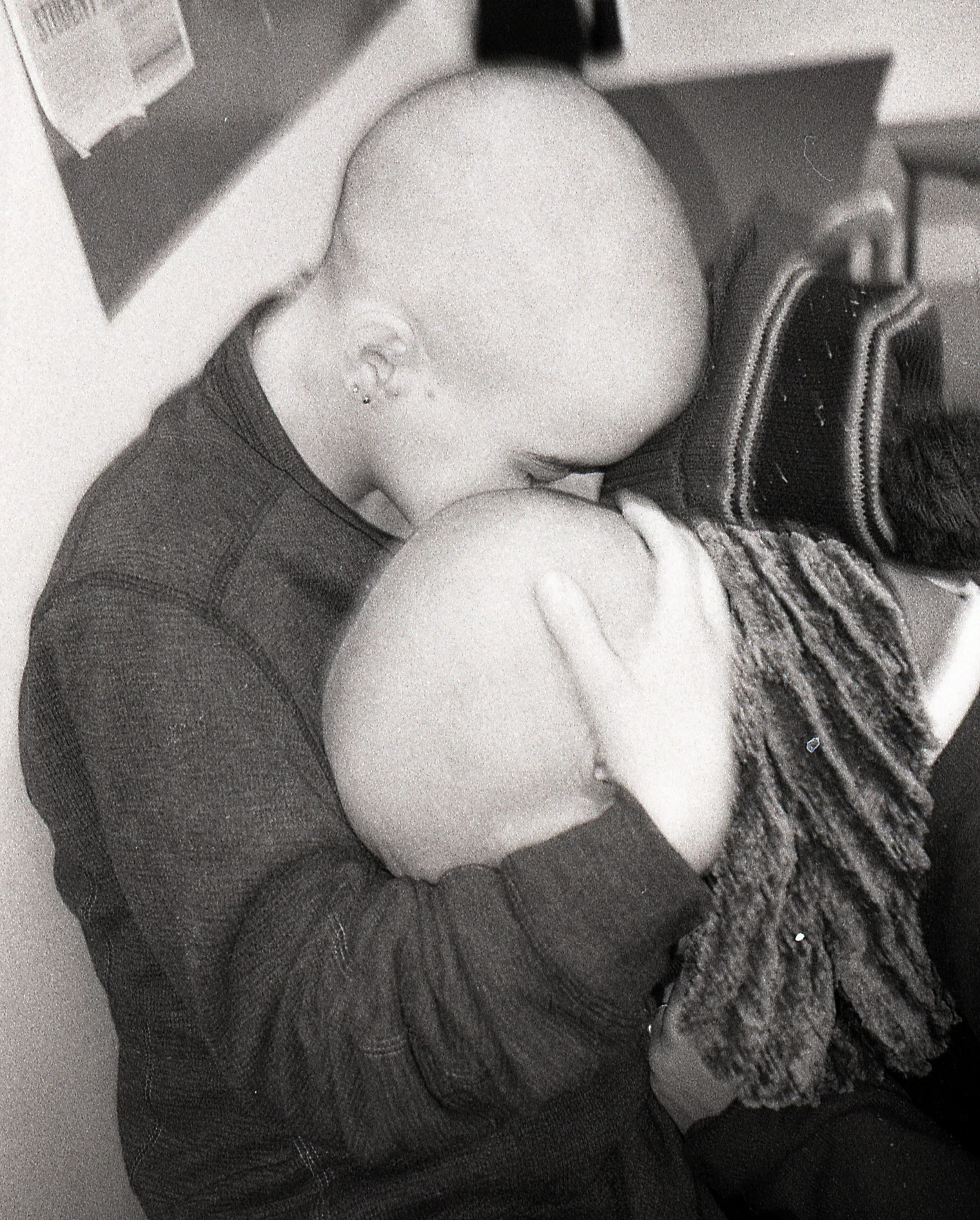 A person with a bald head and earrings holding and kissing a bald child's head, both facing each other.