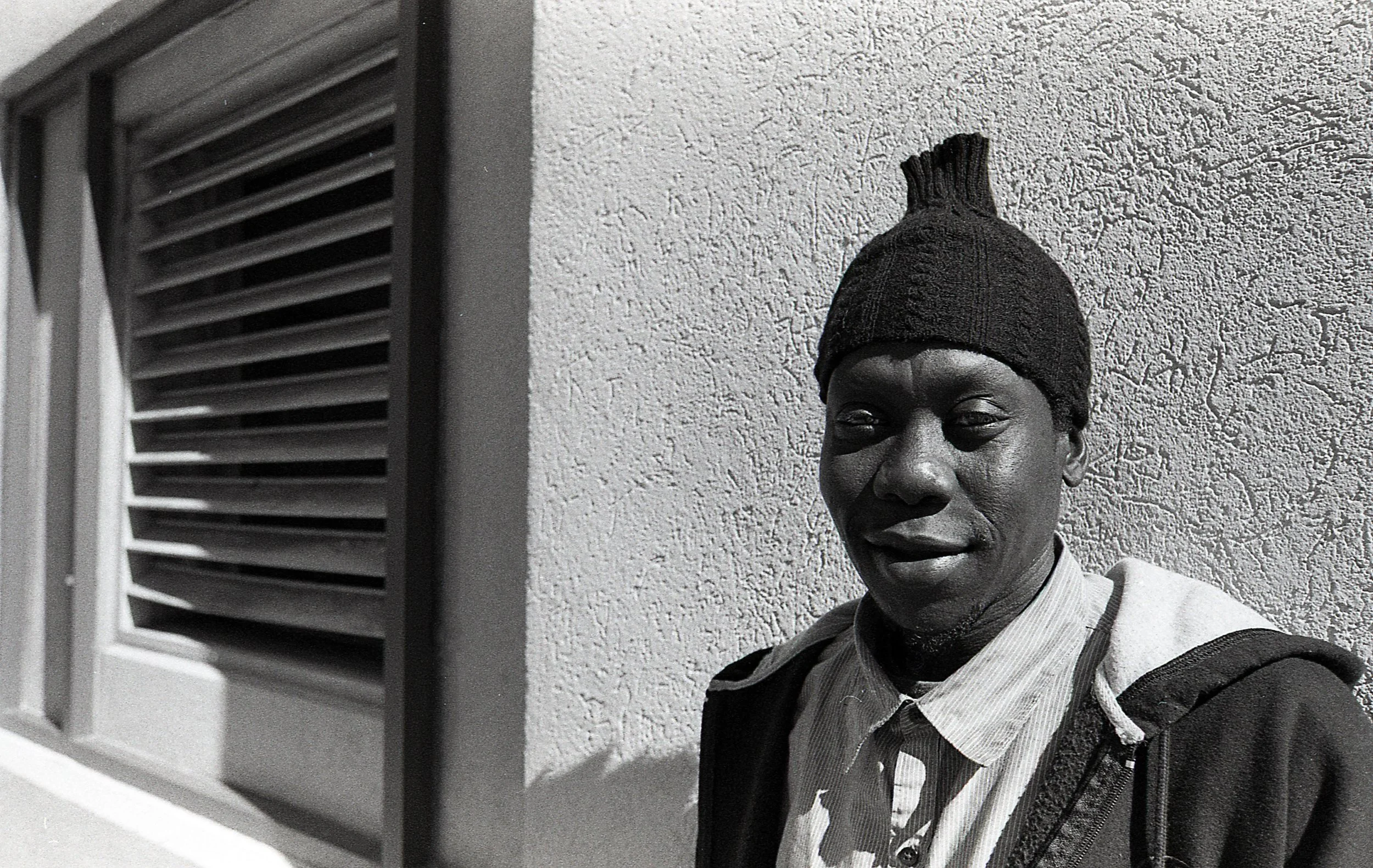 A young man standing outdoors against a textured wall, wearing a beanie hat, a collared shirt, and a jacket, with a window with horizontal blinds nearby.
