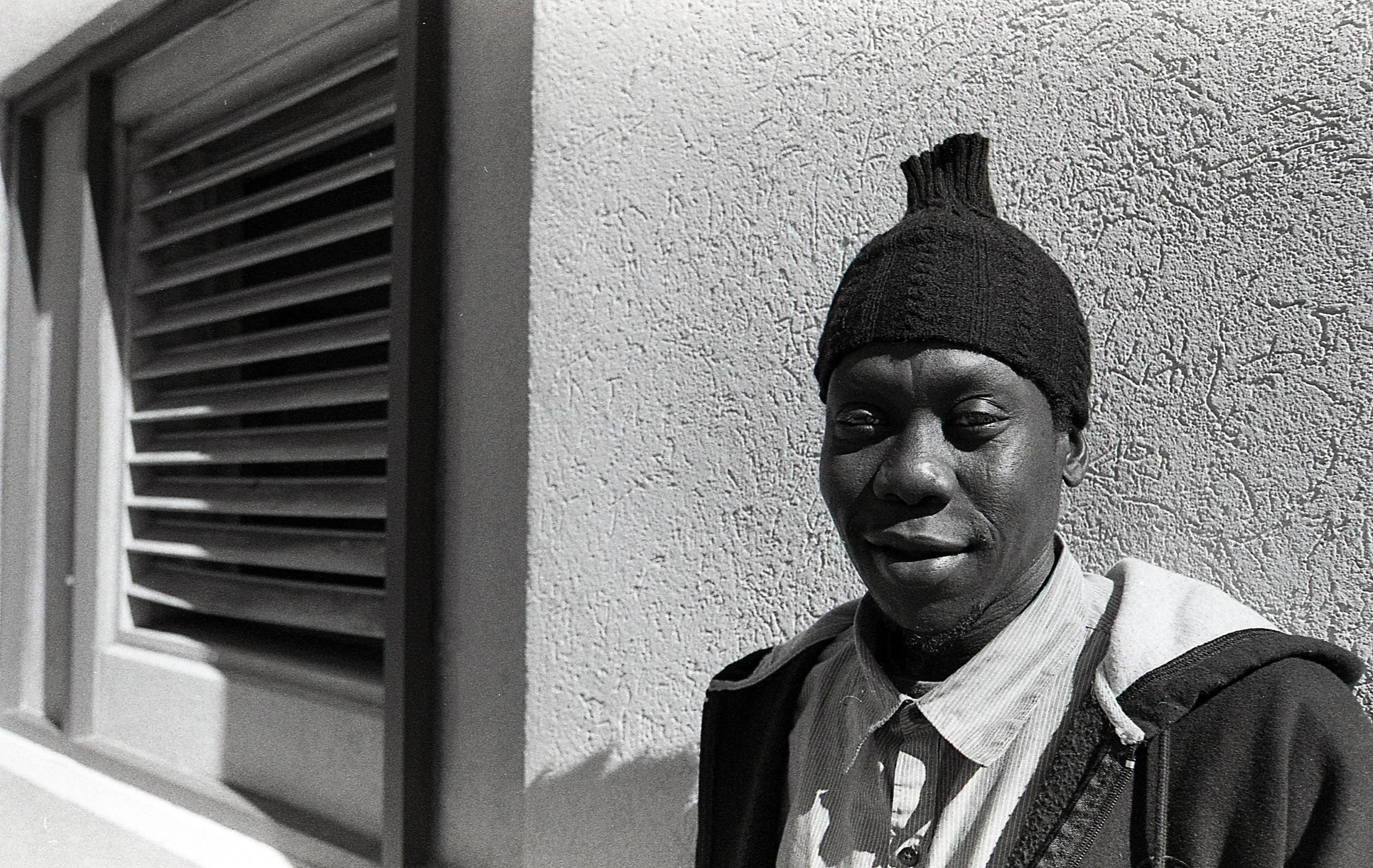 A man with dark skin wearing a knit cap and a hoodie, standing against a textured wall and a window with horizontal blinds.