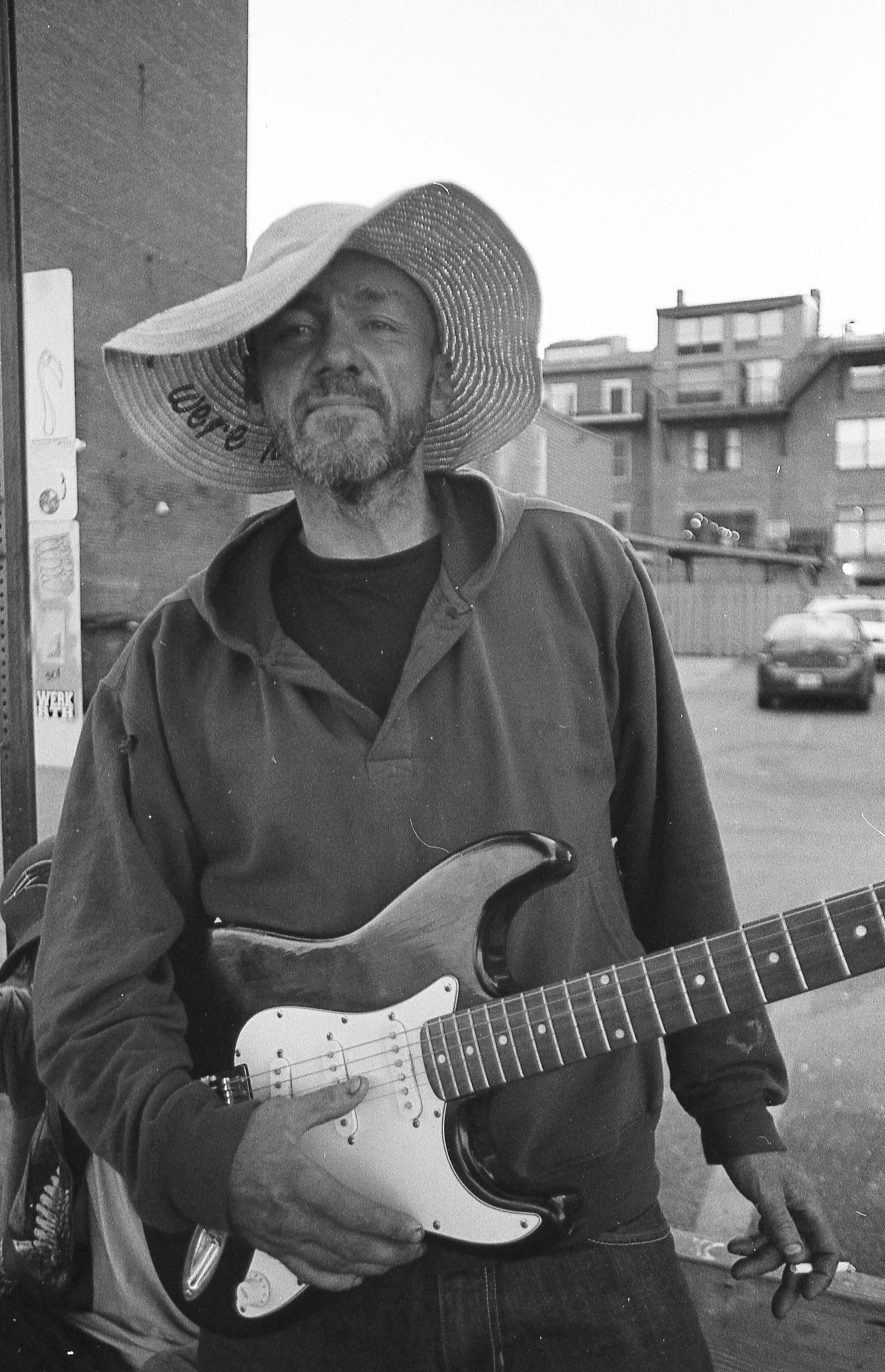 A man wearing a large sun hat and a dark hoodie, holding an electric guitar, standing outdoors in an urban area with buildings and parked cars in the background.
