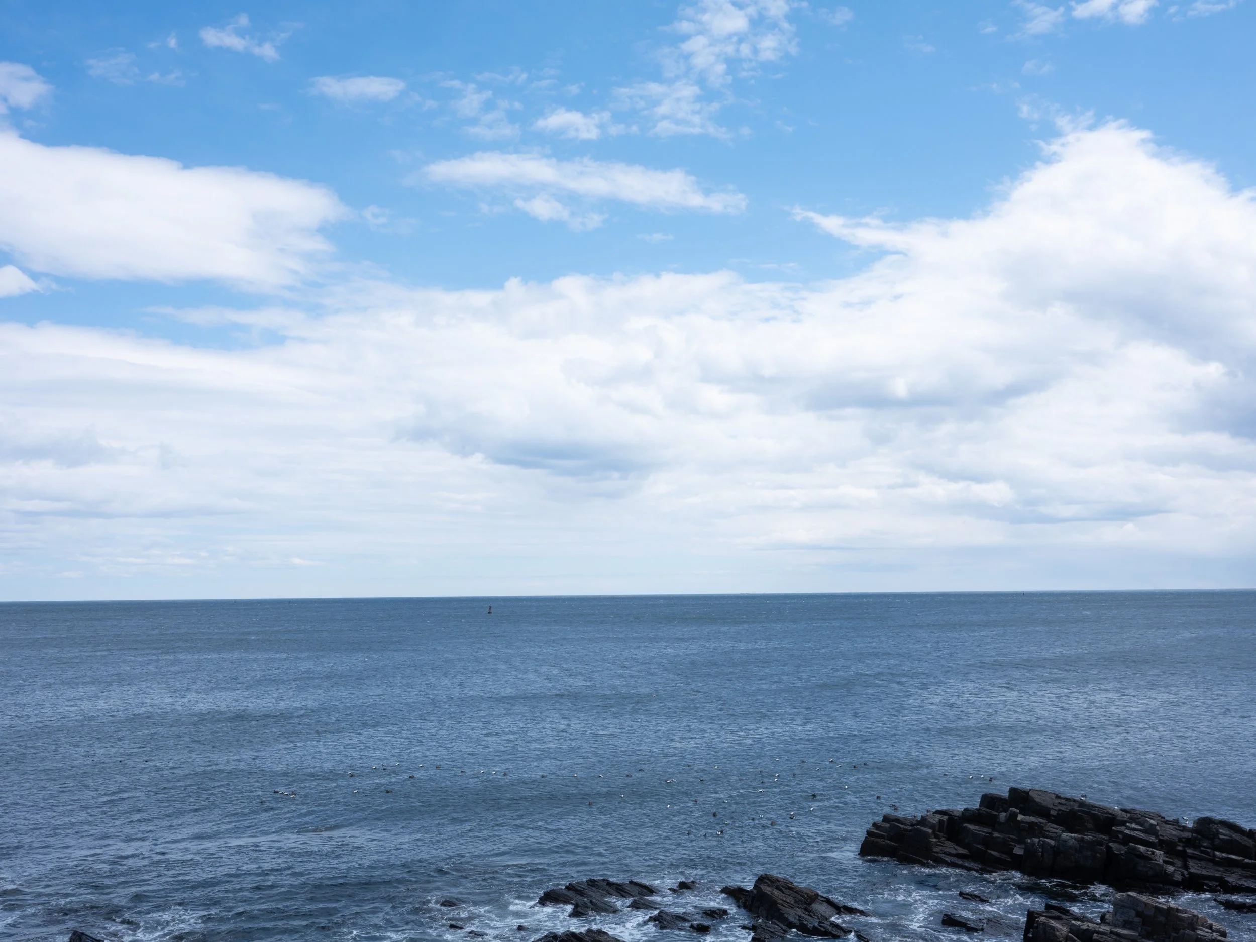 Ocean view with a partly cloudy sky, dark rocks at the shoreline, and distant horizon