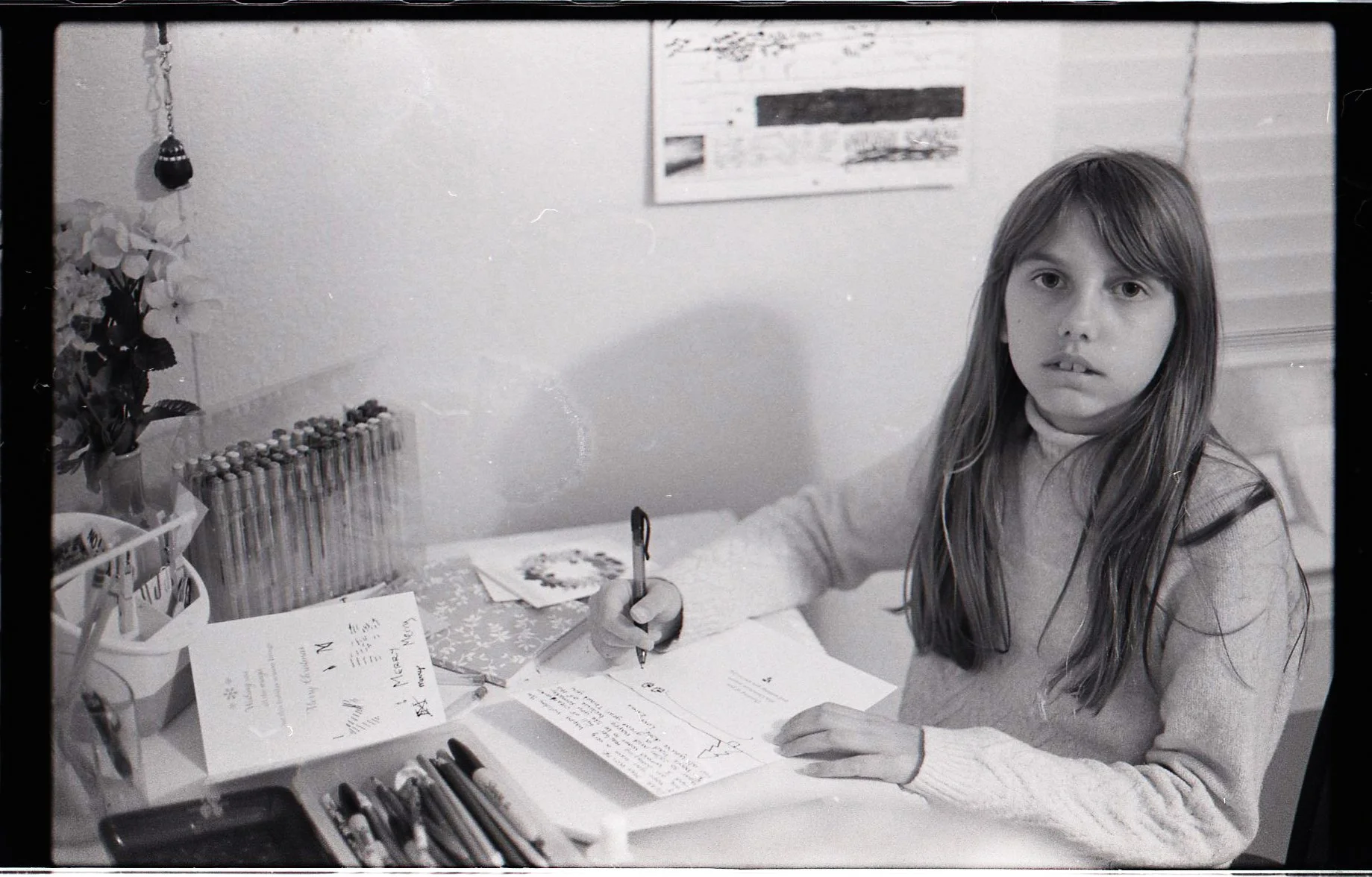 A young girl with long hair writing a letter at a desk with various pens, pencils, and greeting cards. There is a potted plant and a row of colored pencils on the desk, with a wall art piece in the background.