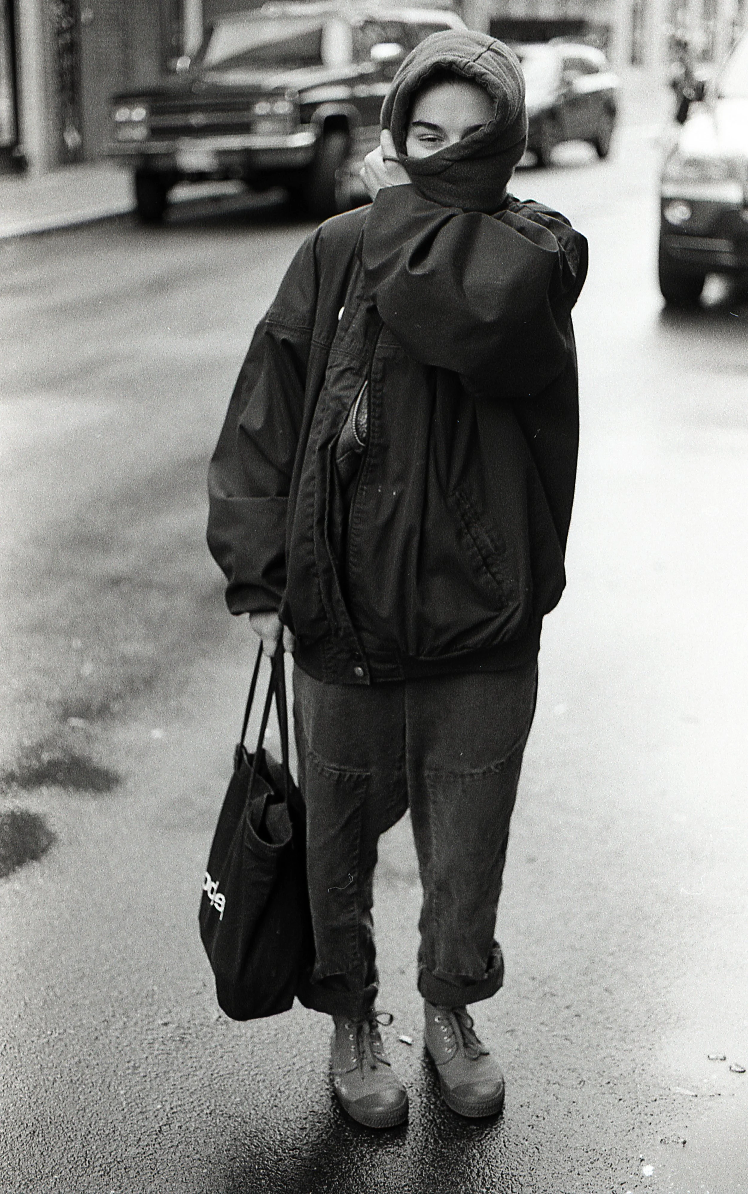 A person standing on a wet street wearing a hoodie, jacket, and jeans with a backpack and carrying a shopping bag, with parked cars in the background.