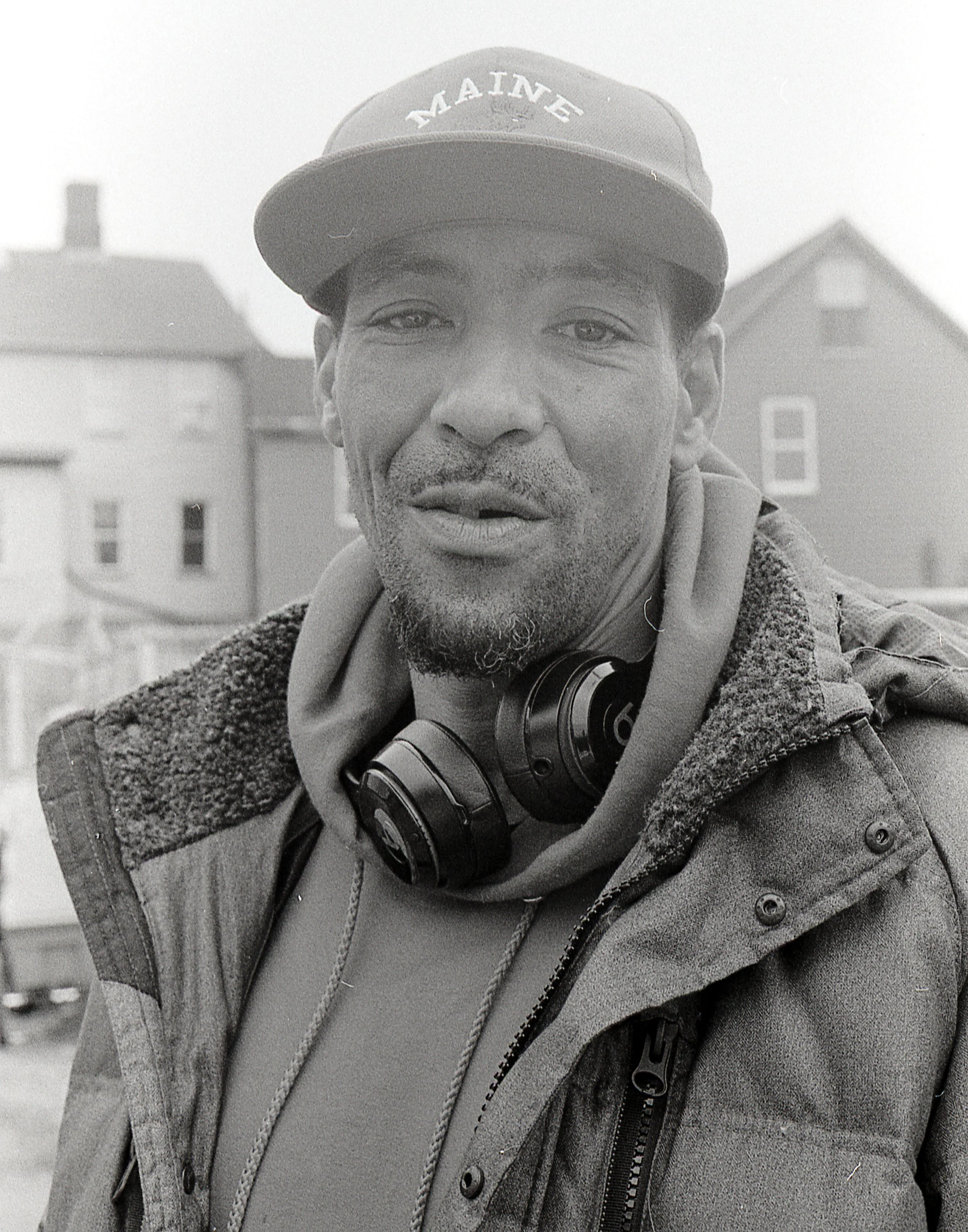 Black and white photo of a man wearing a cap with 'MAINE' on it, a hoodie, and a jacket, with headphones around his neck, standing outdoors with houses in the background.