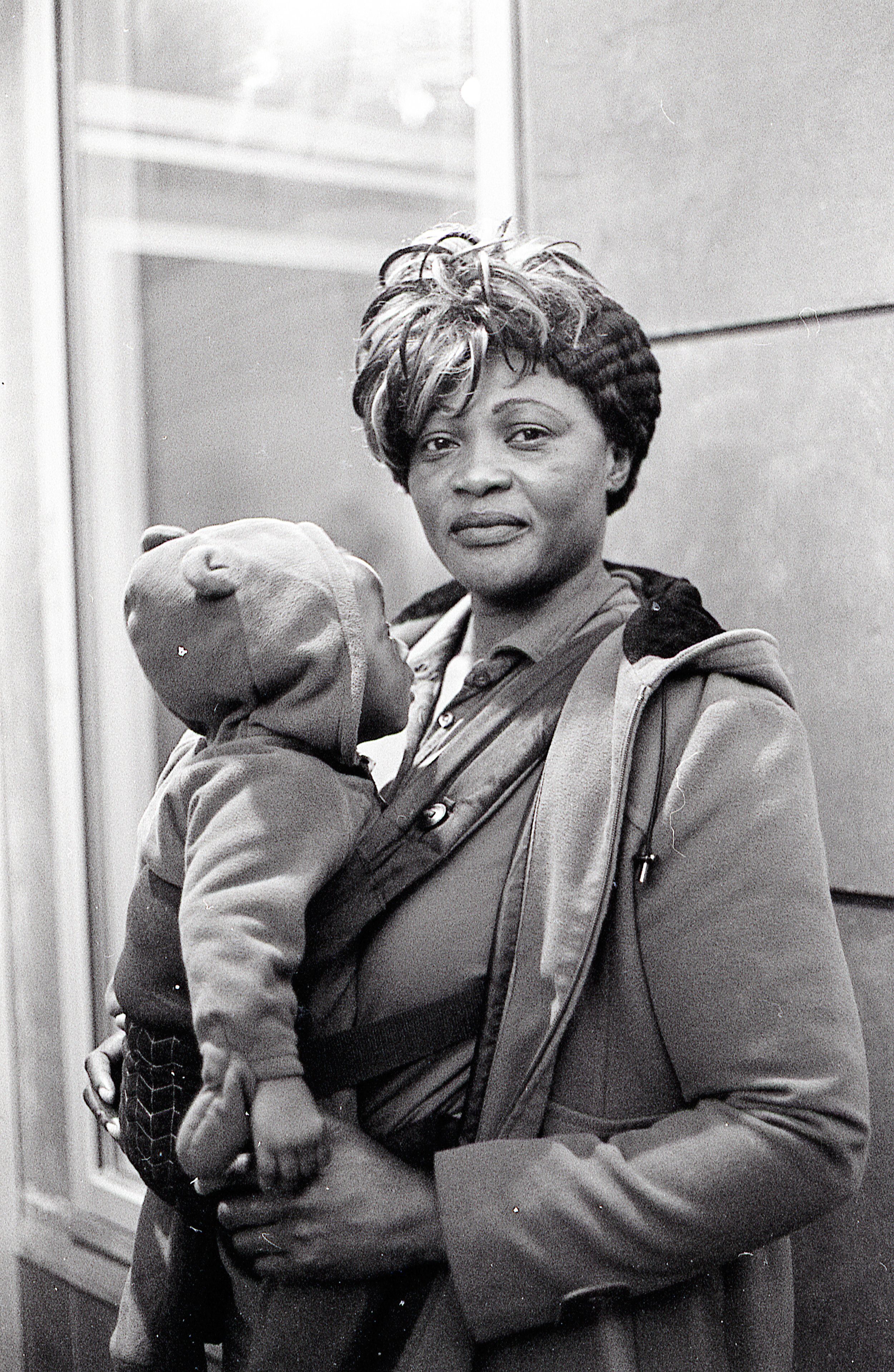 A woman with short, styled hair holds a small child wearing a hat with animal ears. The woman is looking directly at the camera and is dressed in a jacket, while the child is resting their head on her chest.