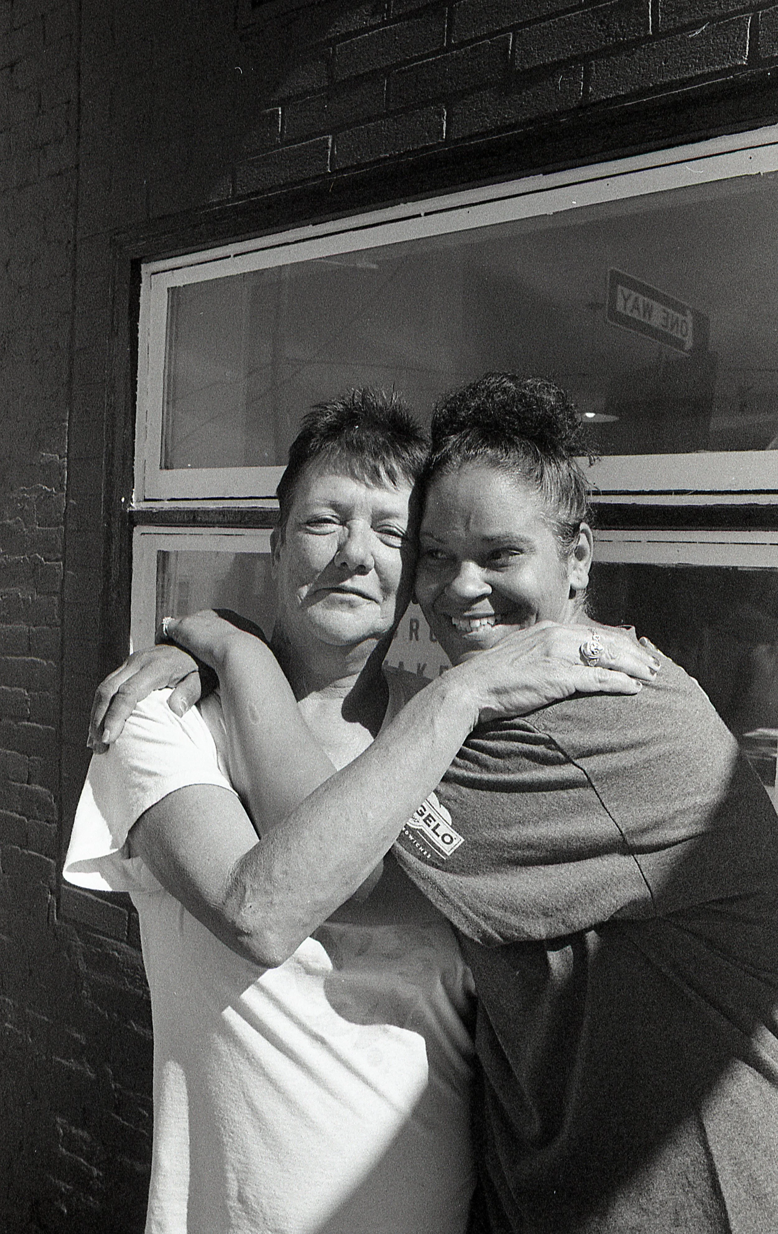 Two women hugging, standing in front of a brick wall and window, smiling.