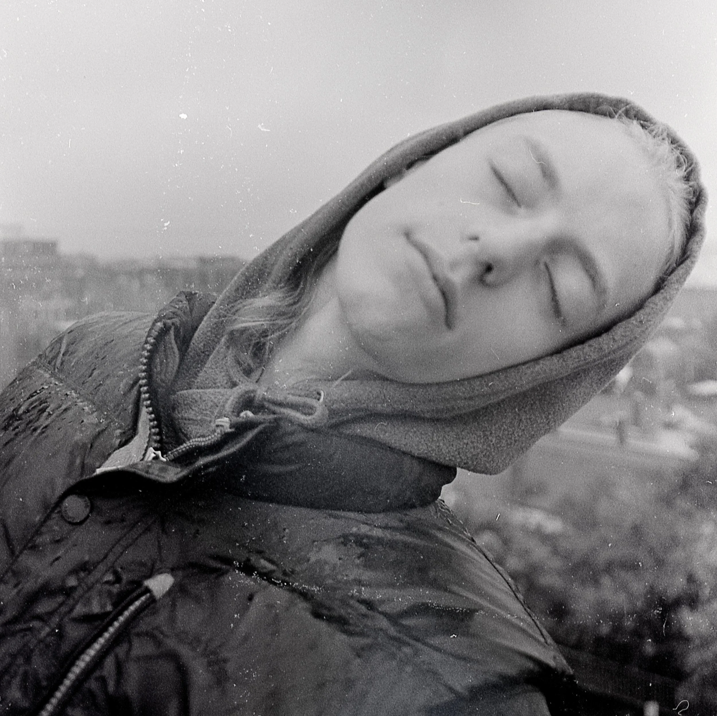 A person with closed eyes wearing a hoodie and a jacket outdoors in black and white.