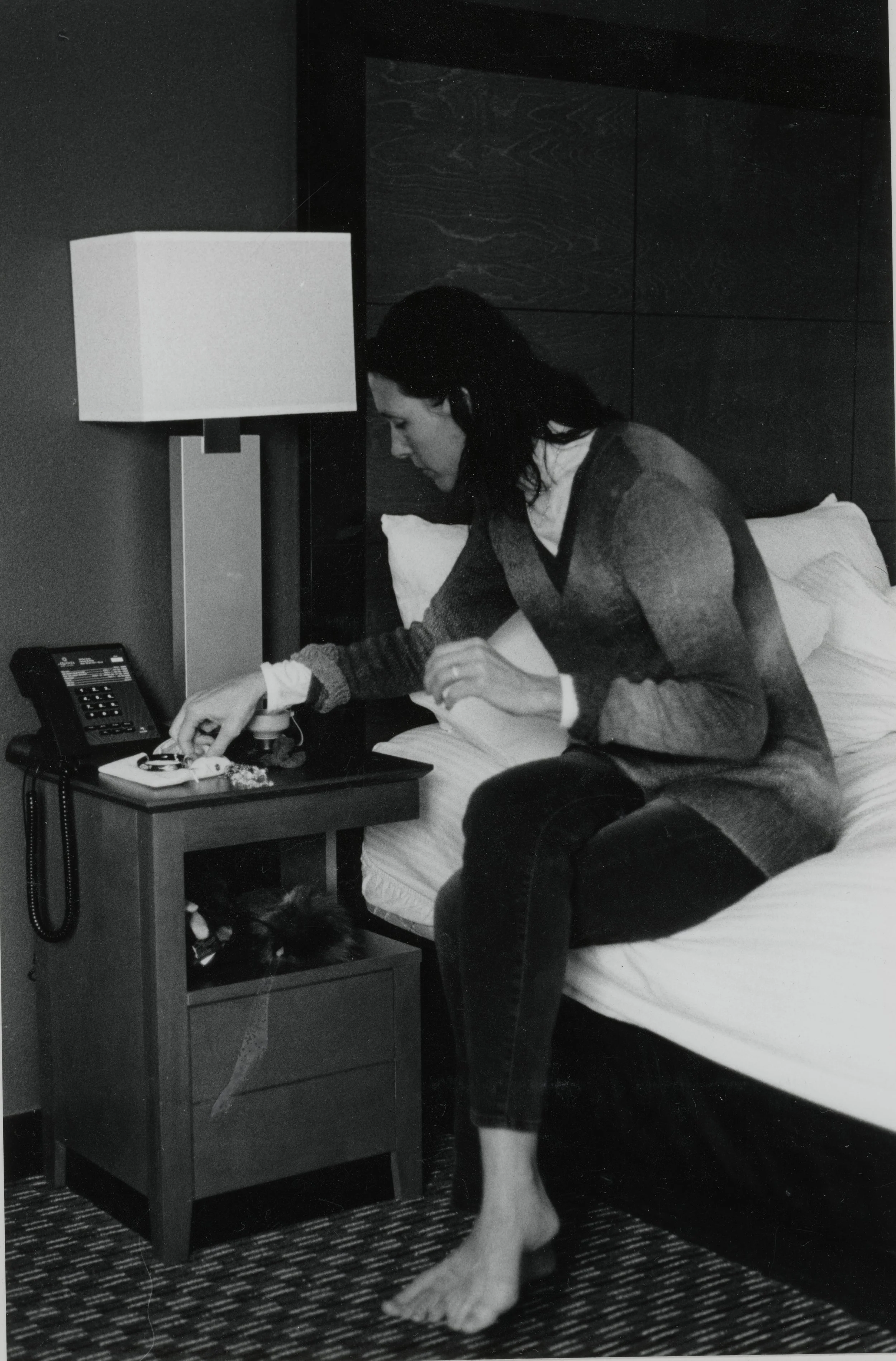 A woman sitting on a hotel bed, reaching for a phone on the nightstand, with a cat under the nightstand, in a black-and-white photo.