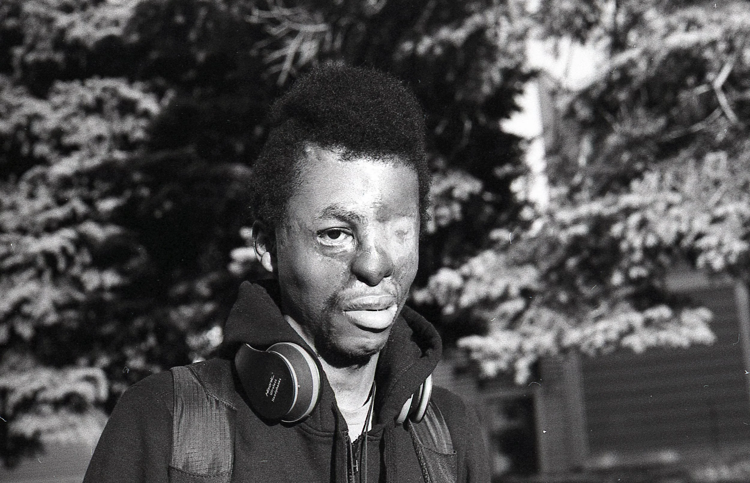 A young man with short afro hairstyle wearing headphones and a dark hoodie, standing outdoors in front of a snow-covered tree at night.