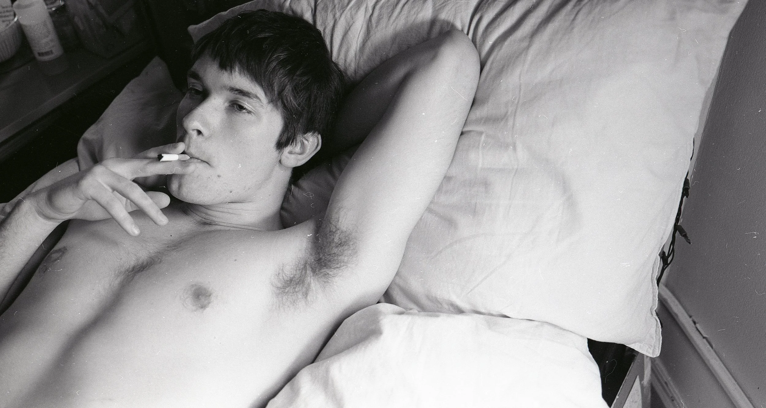 A young man with dark hair lies shirtless in bed, smoking a cigarette, with his head resting on a pillow and his arm behind his head.