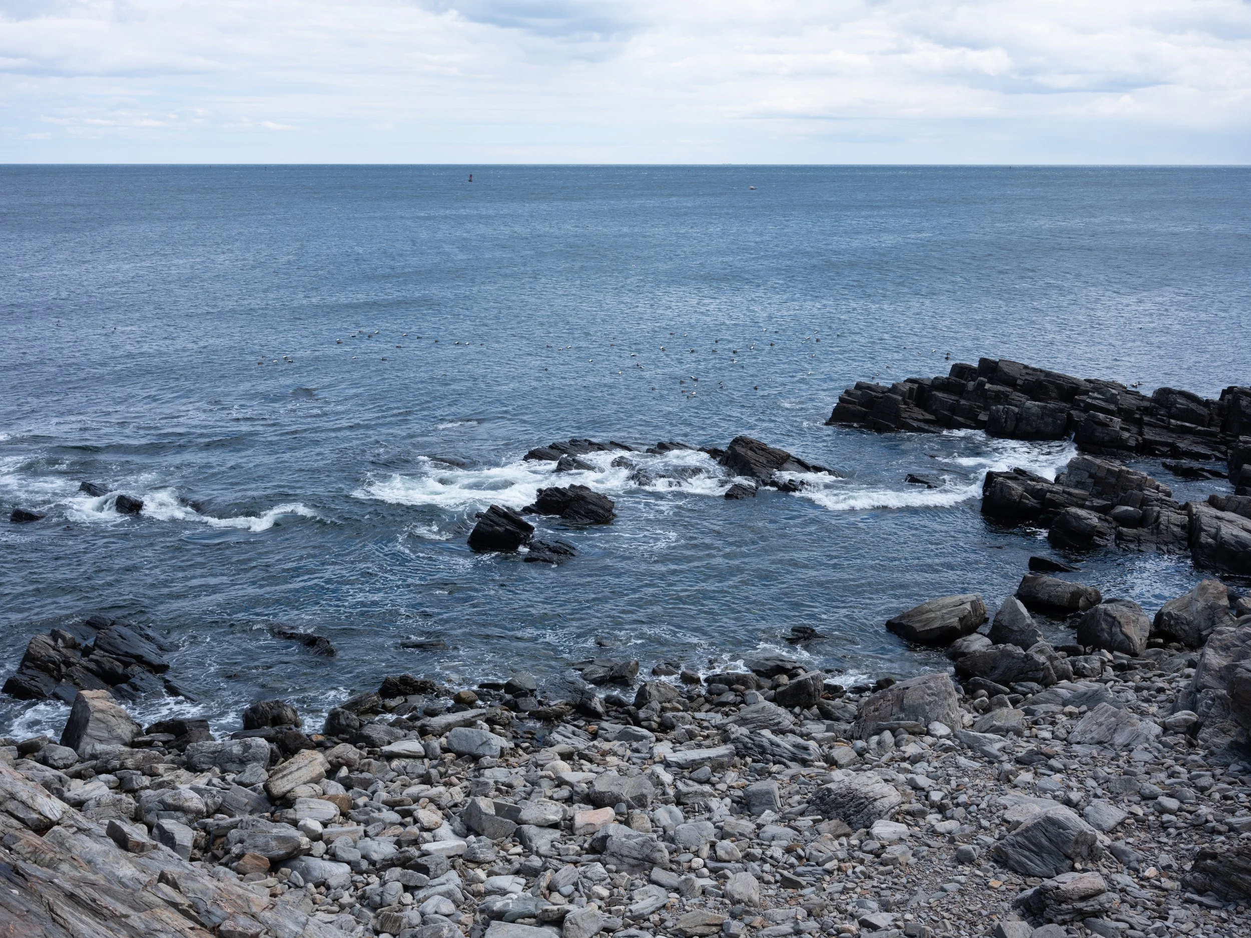 Rocky shoreline with waves crashing against rocks and a calm ocean extending to the horizon, under a partly cloudy sky.