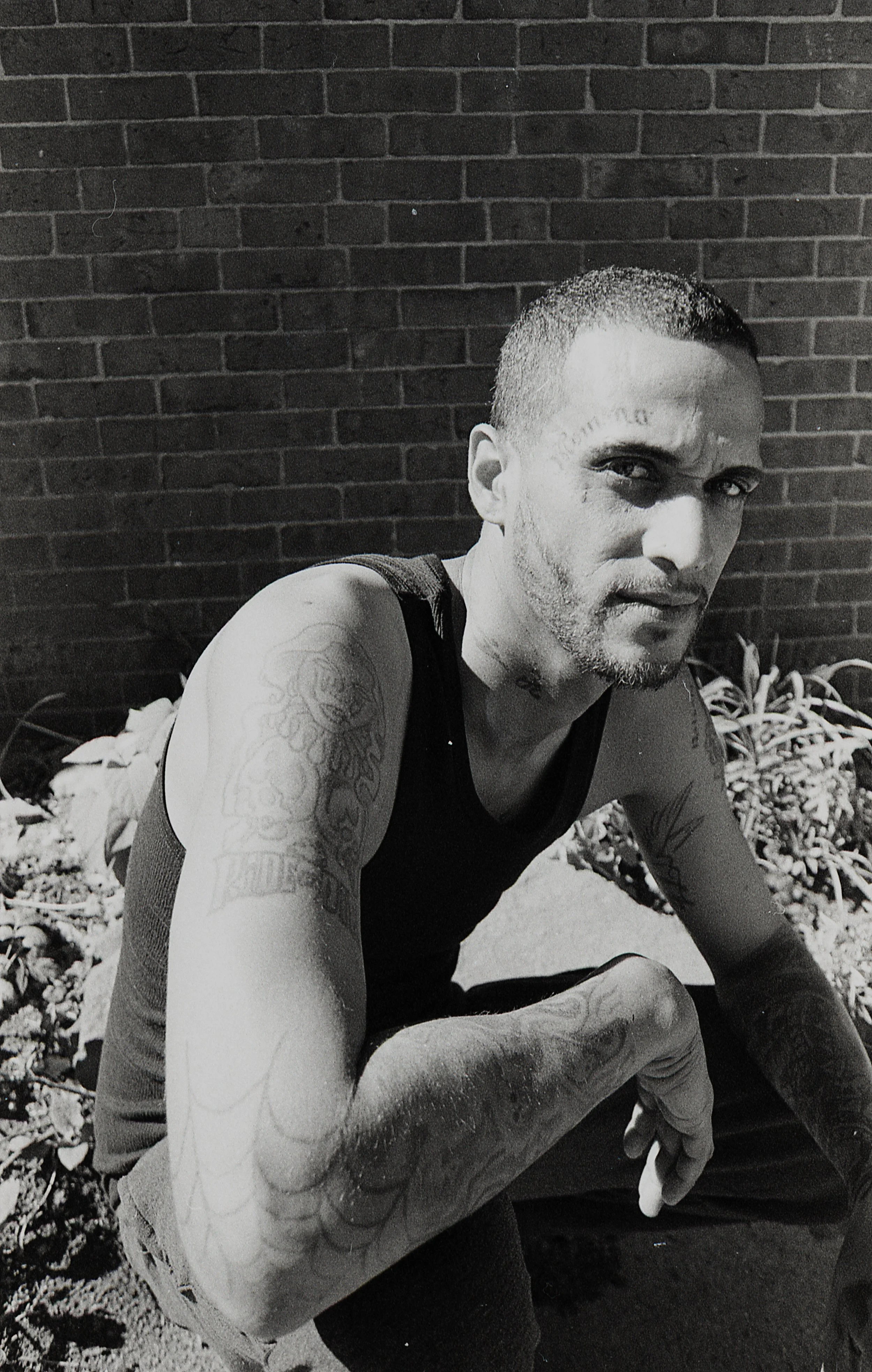Black and white photo of a man with tattoos on his arm, wearing a sleeveless shirt, sitting outdoors against a brick wall, with weeds in the background.