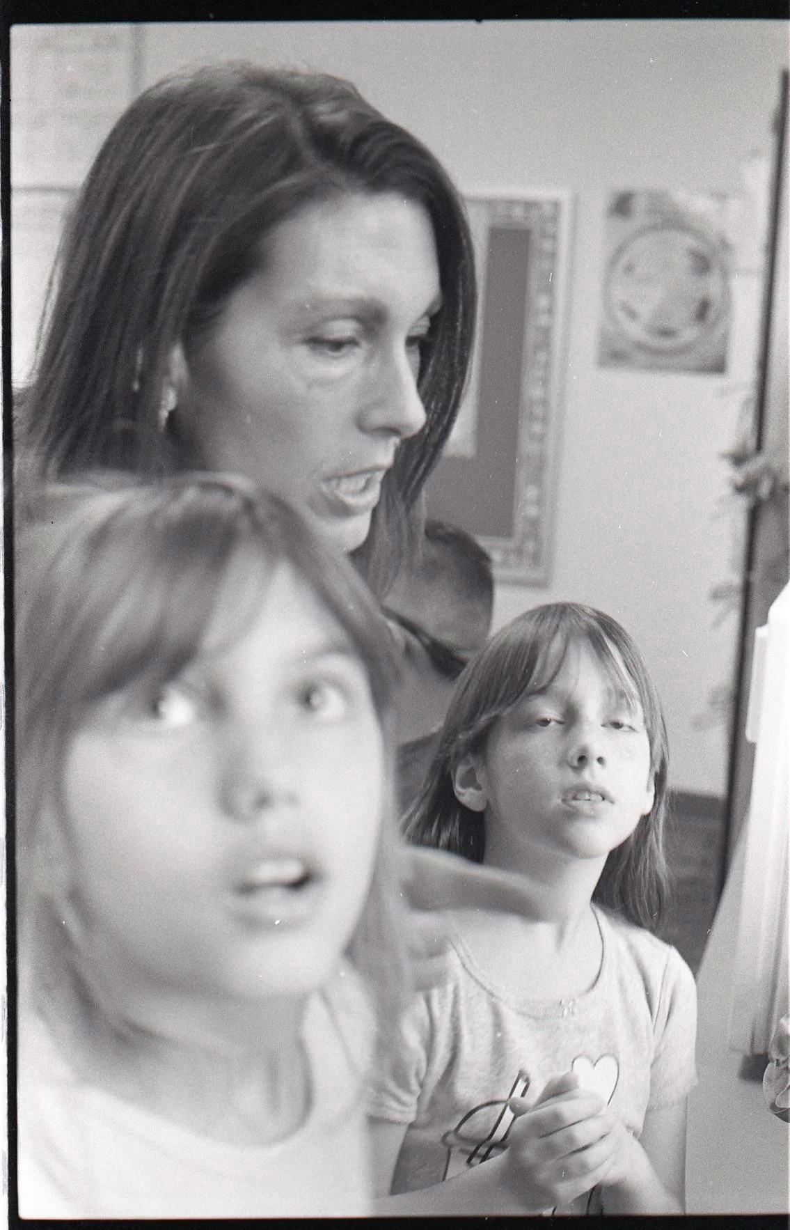 Three girls in a room, with two of them faced slightly away from the camera. The girl in front has short hair and an expression of surprise. The girl in the background has long hair and is looking at the camera with a neutral expression.