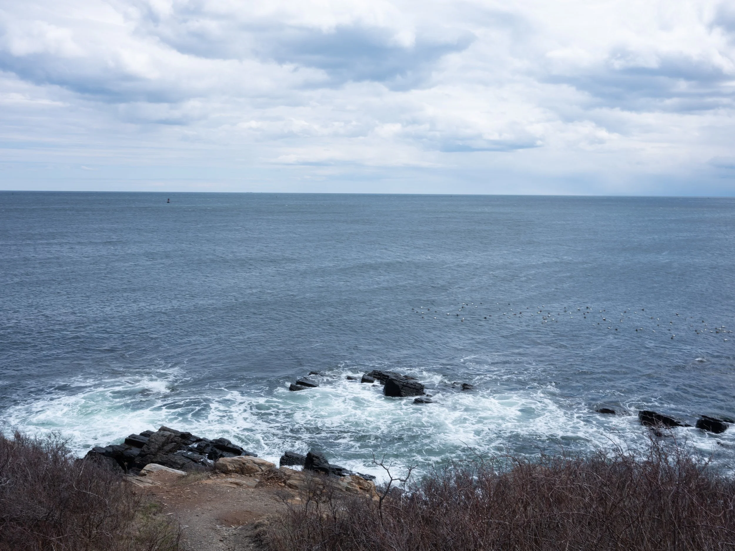 A view of the ocean with rocks and a rocky shoreline in the foreground, and a cloudy sky above.
