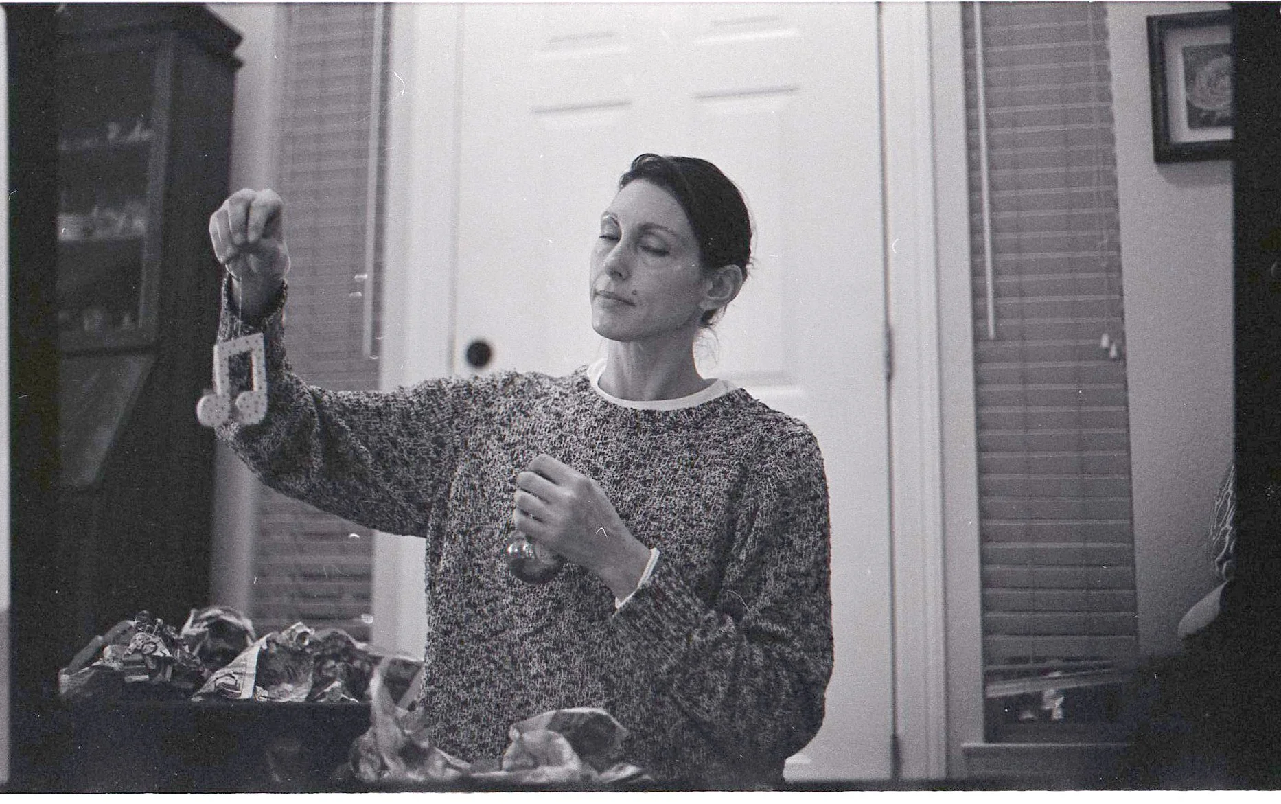 A woman with short dark hair wearing a patterned sweater stands in a kitchen, holding a small object in her left hand and a keychain with a photo in her right hand, looking at the keychain. There are cabinets, a doorway, and a bag or box filled with 