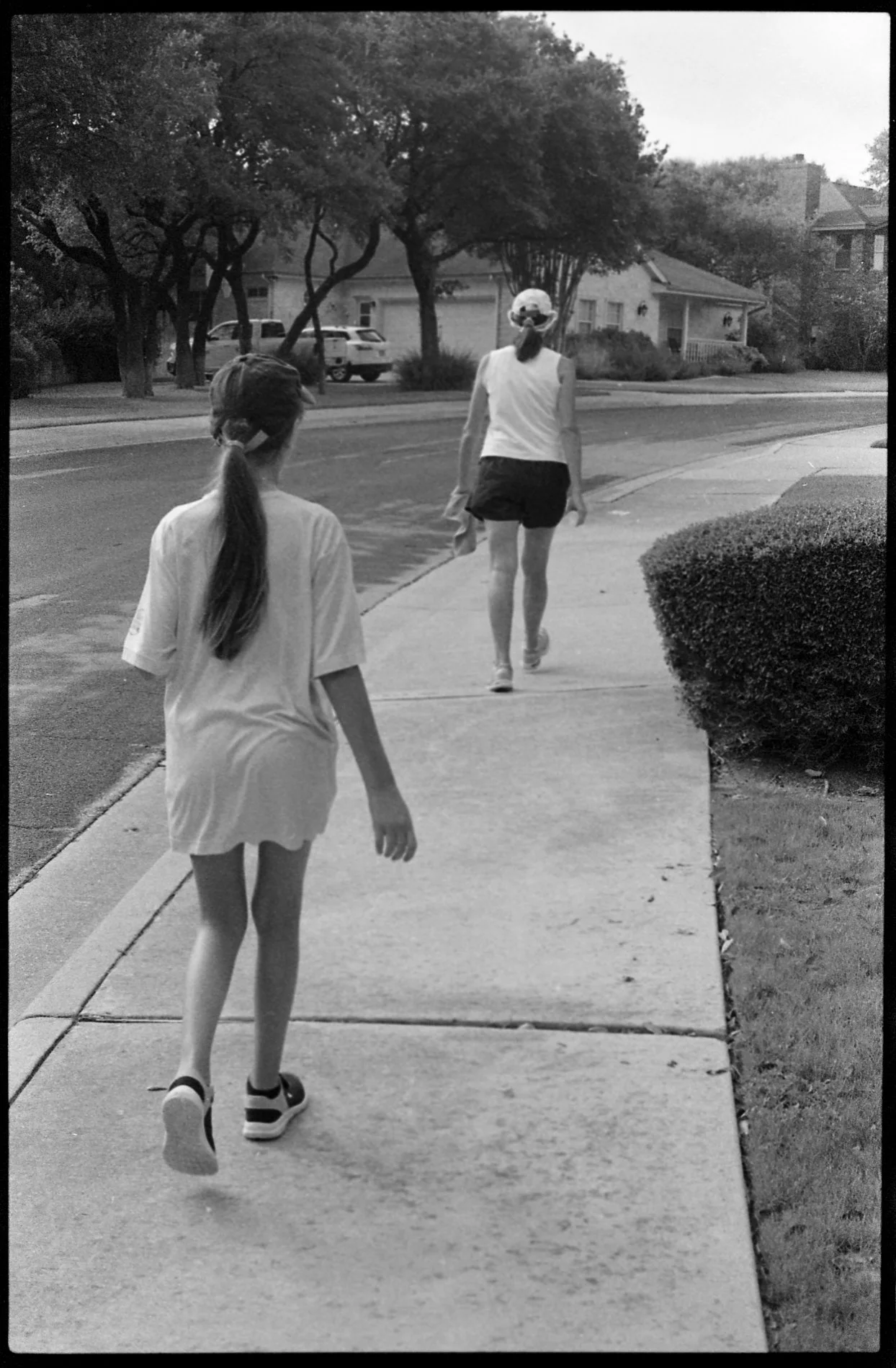 A girl with a ponytail wearing a loose t-shirt and sneakers walking on the sidewalk, approaching a woman wearing a hat and a tank top, carrying what appears to be a towel or cloth, as they walk away from the camera.