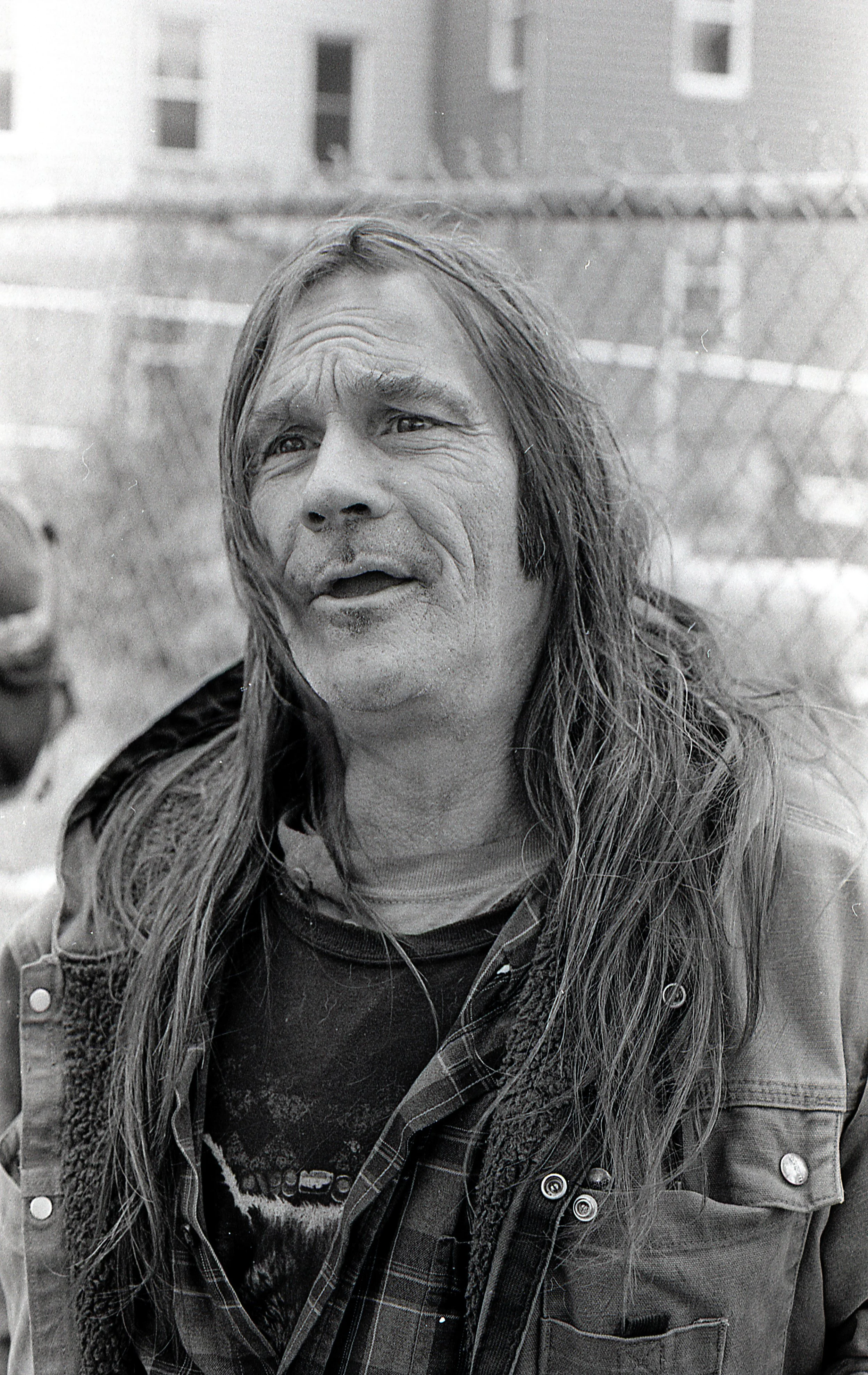 Black and white photo of a man with long hair, wearing a jacket and plaid shirt, outdoors near a chain-link fence and buildings.