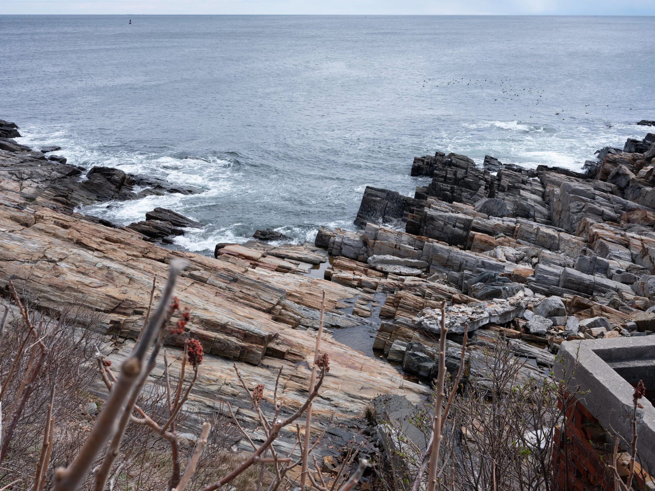 Rocky shoreline by the ocean with waves crashing against the rocks and a small boat far in the distance.