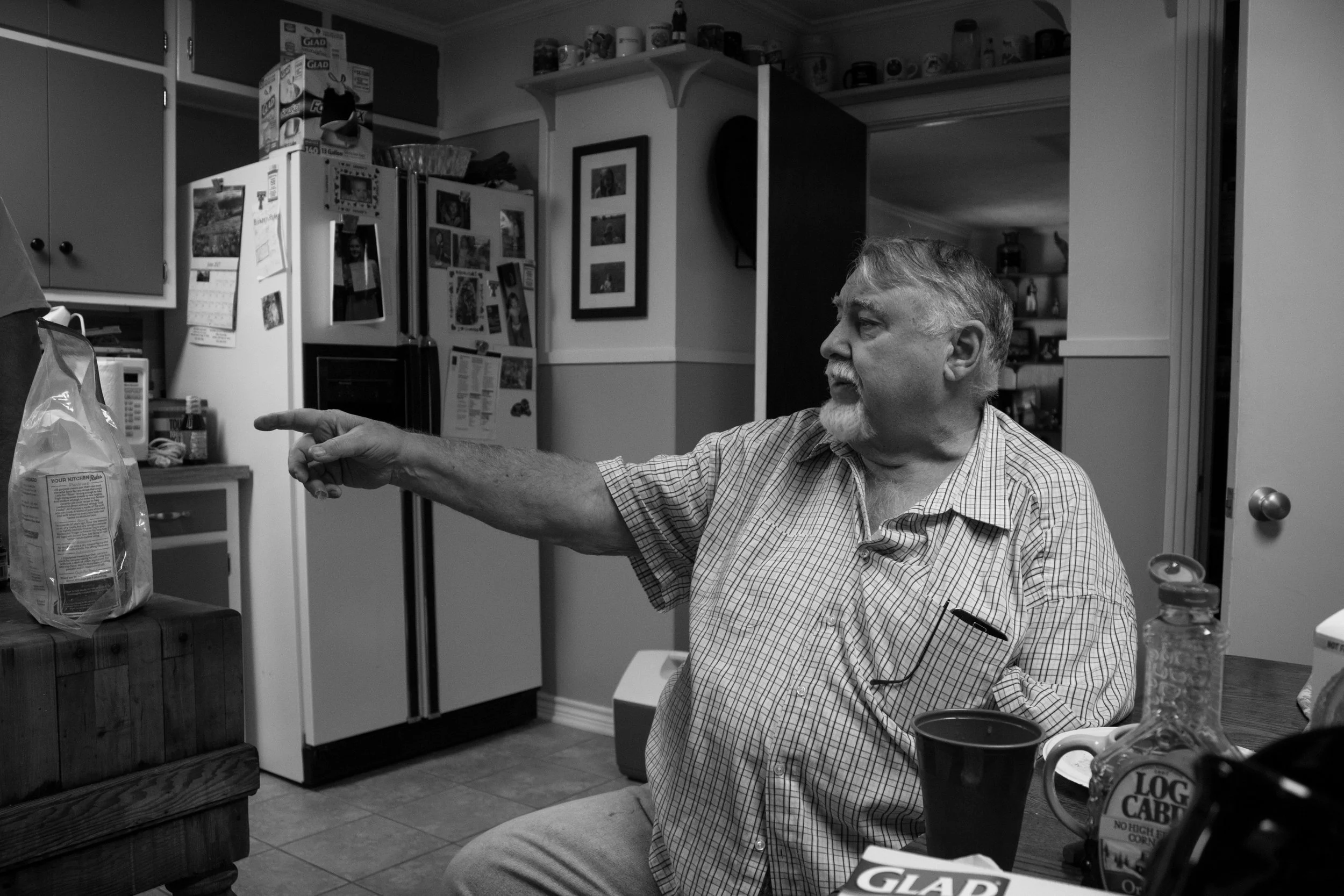 An older man with gray hair and beard sitting at a kitchen counter, pointing to a bag of food with his right hand, with kitchen appliances and photo frames visible in the background.