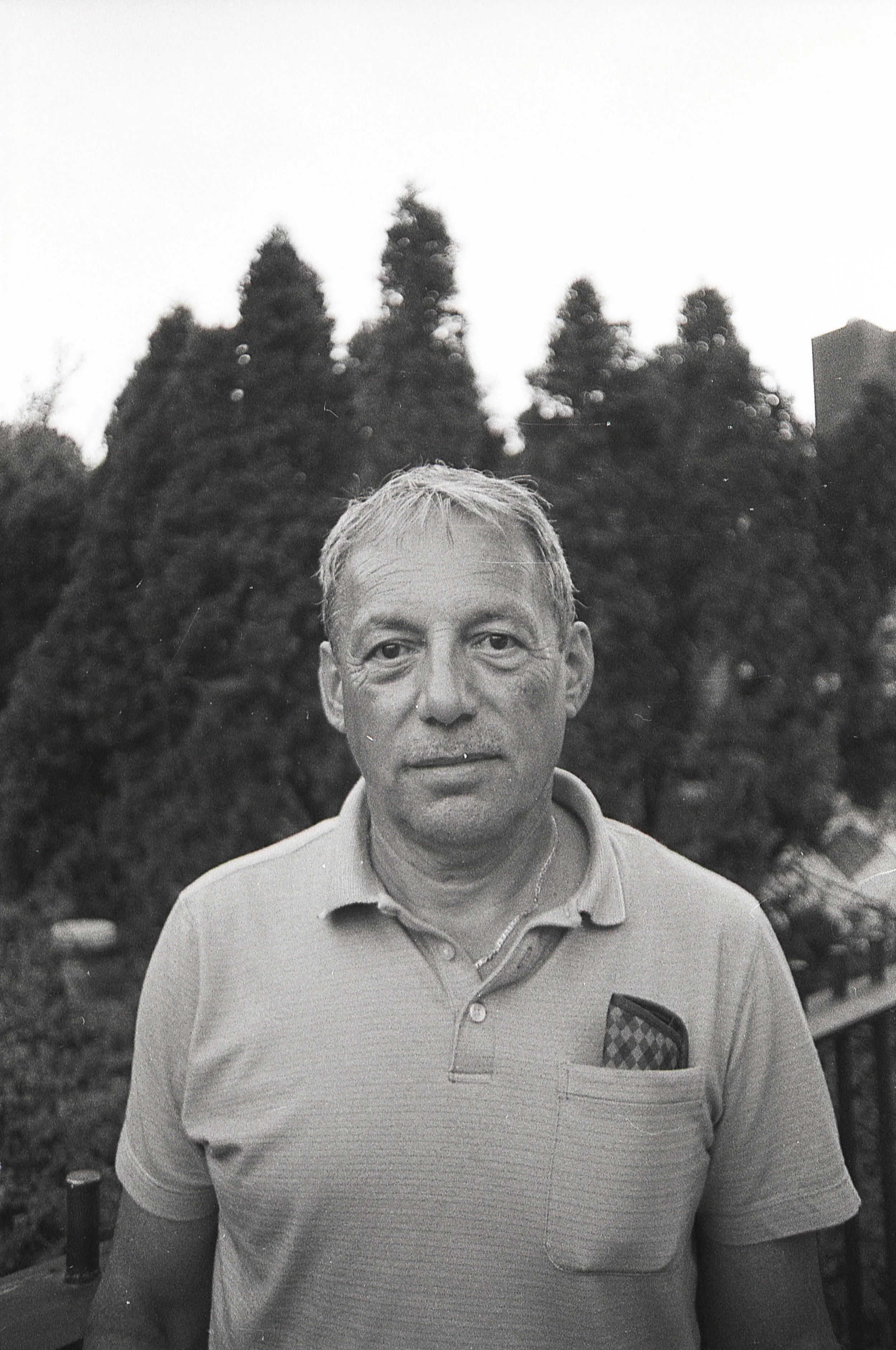 Black and white photograph of a middle-aged man standing outdoors with trees in the background. The man has short, light-colored hair and is wearing a polo shirt with a pocket containing a checkered cloth. He is looking directly at the camera with a 