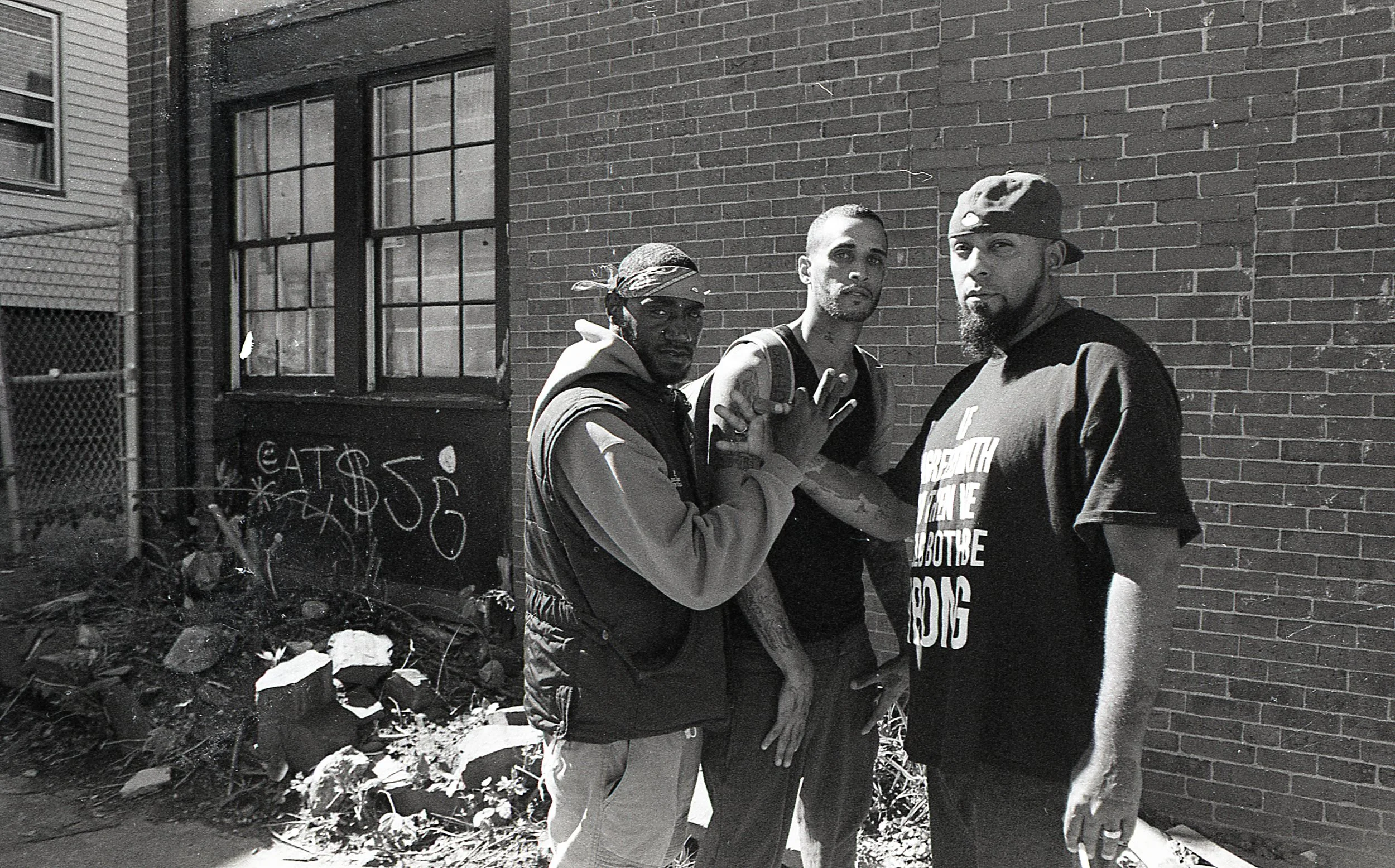 Three men standing against a brick wall on an urban street, with graffiti and debris on the ground.