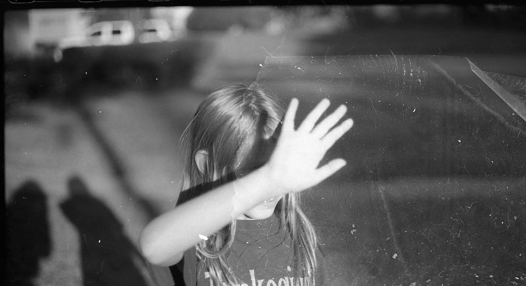 A young girl with long hair holding her hand up against a glass window, blocking her face, with a background of cars and pavement in black and white.