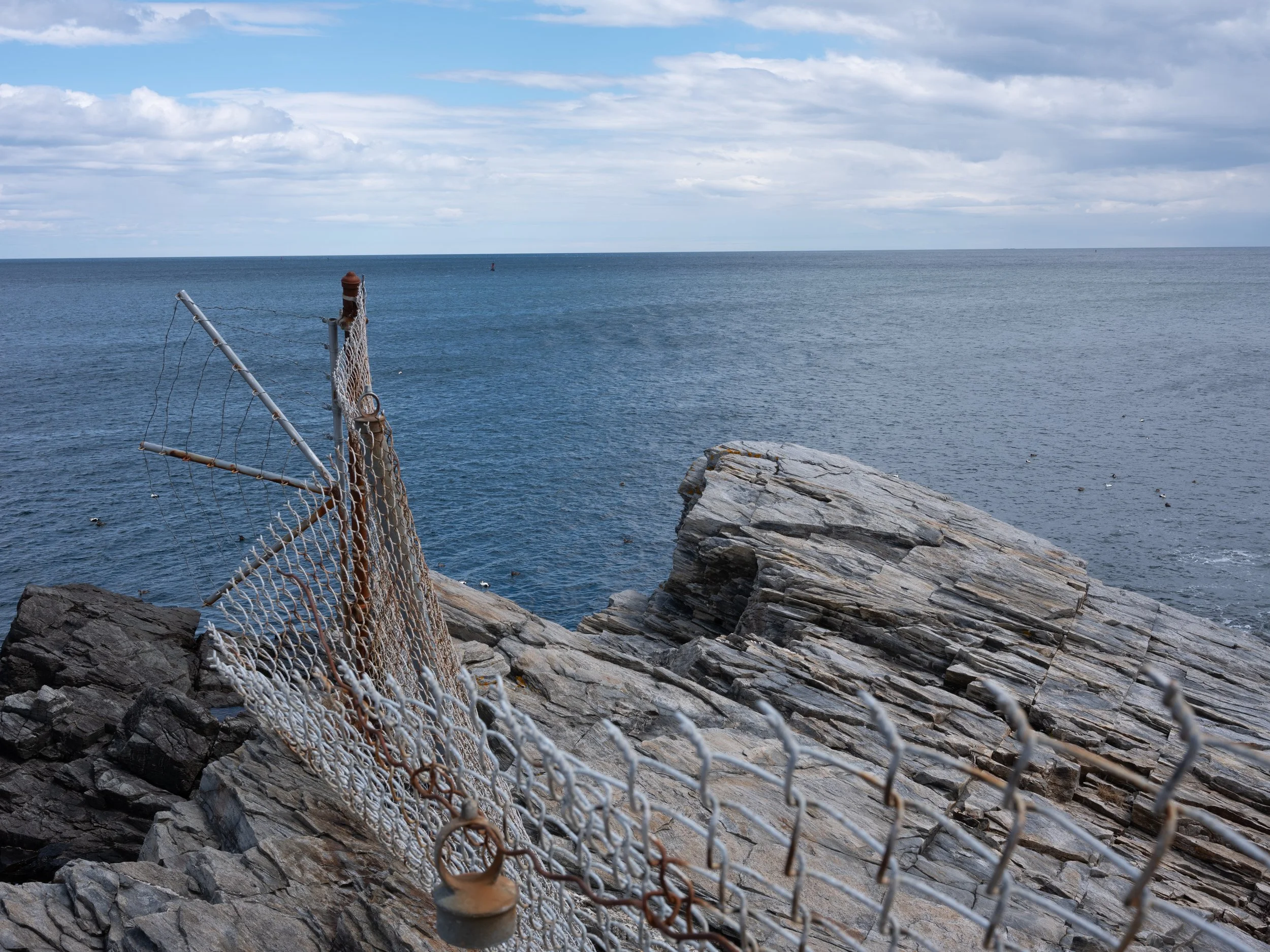 A metal chain-link fence with a rusty lock in the foreground, rocky coastline with large rocks and the ocean in the background under a cloudy sky.
