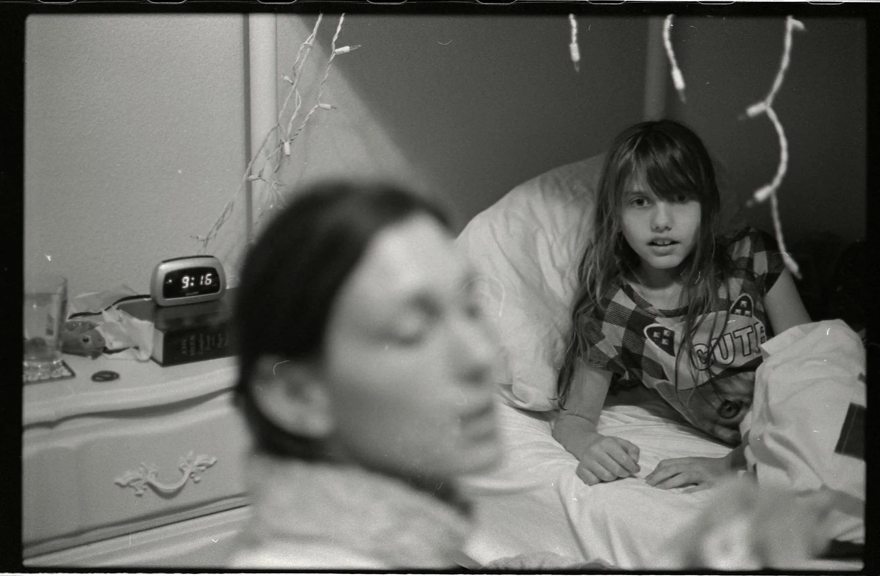A young girl with long hair lying on a bed, looking towards the camera with a surprised or curious expression. In the foreground, a woman with glasses, out of focus, appears to be talking or singing. The bedside table has a clock showing 9:16, a glas
