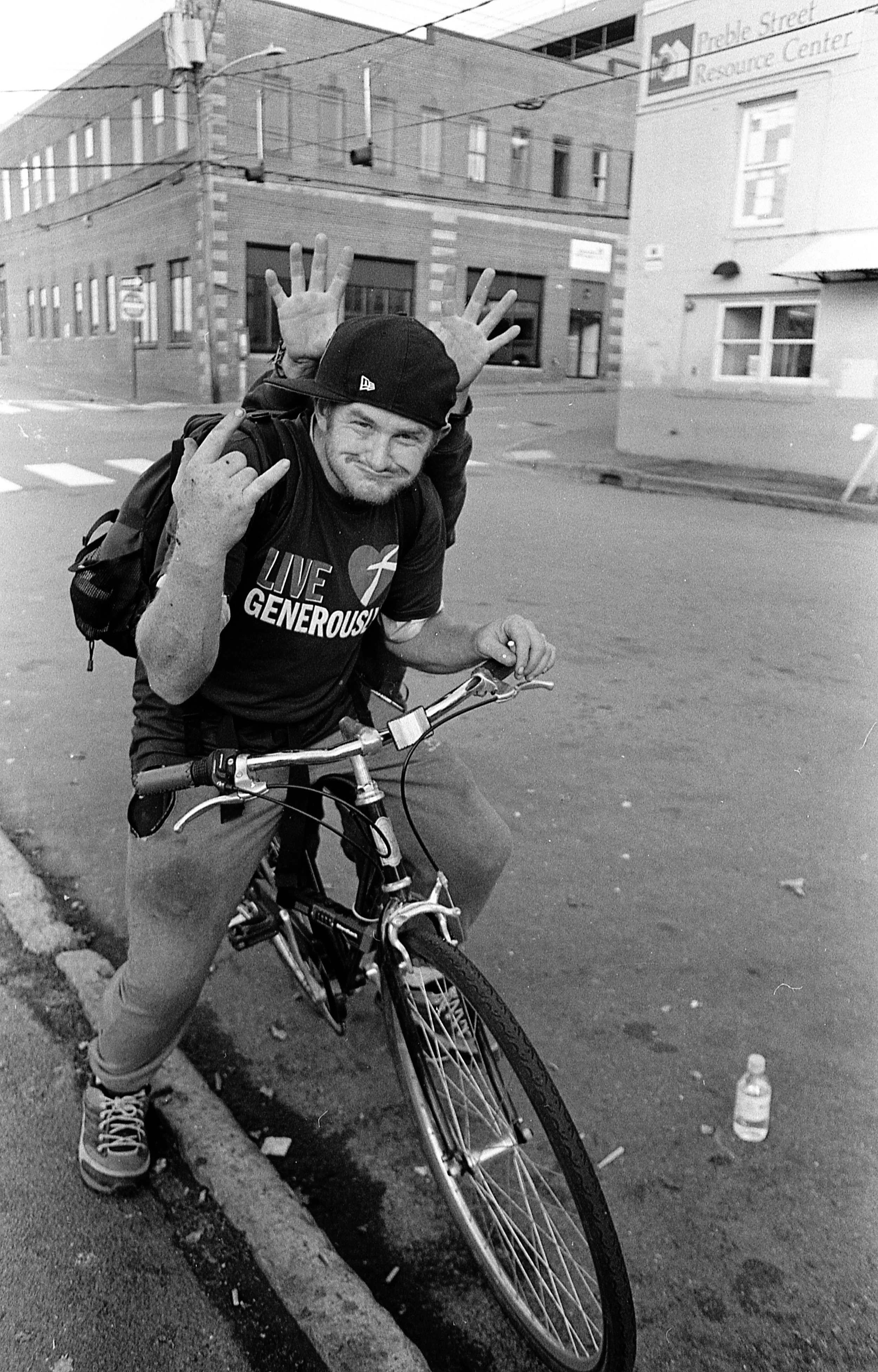 A man with a backpack, wearing a hat and sneakers, is standing on a bicycle on a city street. He makes a hand gesture with his left hand and has a playful expression. There is a water bottle on the ground nearby.