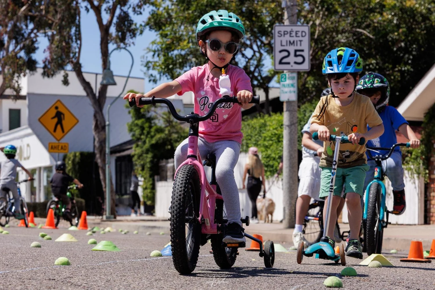 A smiling girl in a red helmet rides a pink bike with training wheels on a sunny day