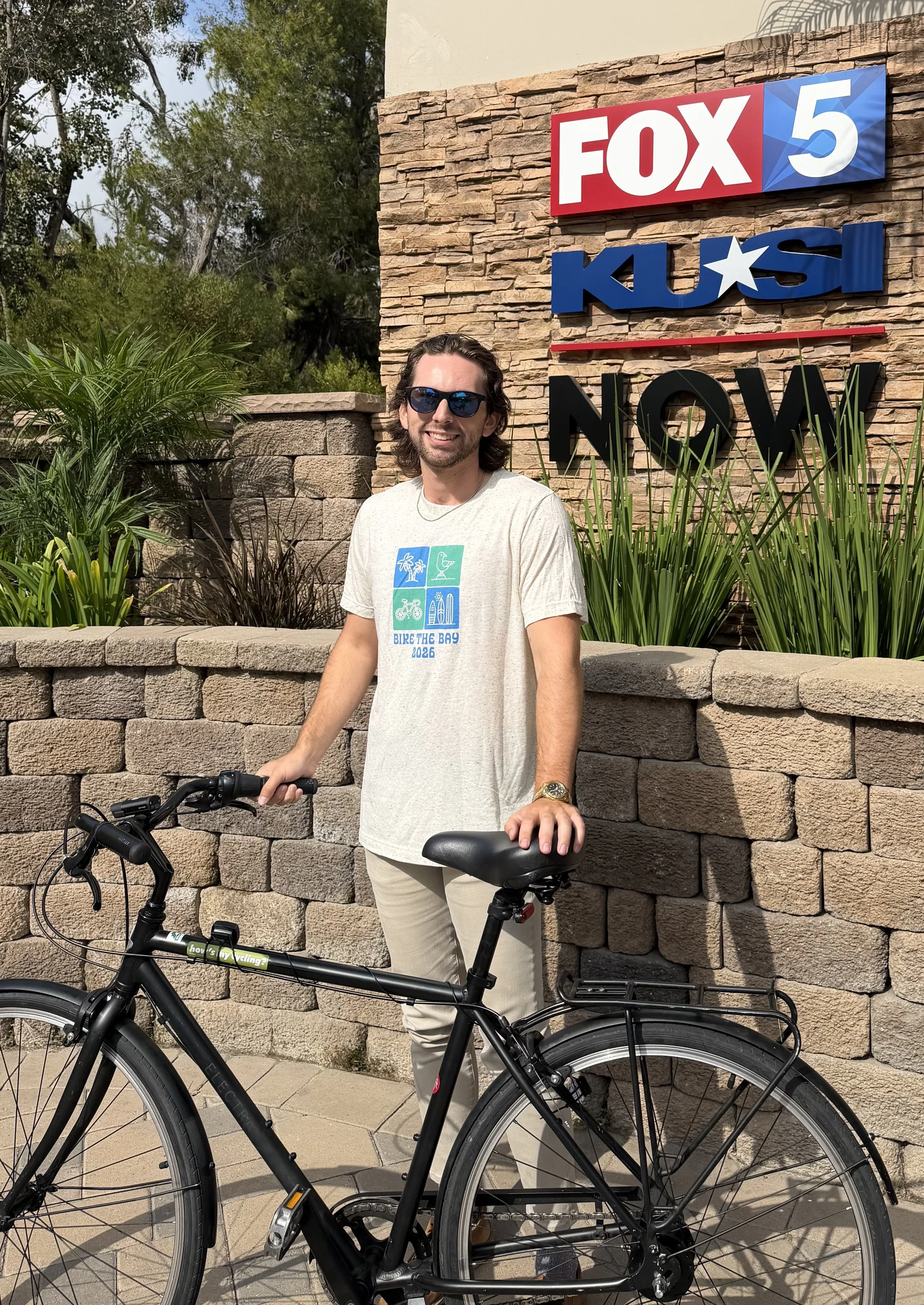 Man wearing a blue bicycle helmet and green T-shirt that says 'LIFE BY BIKE,' sitting on a bicycle on a city sidewalk with crosswalk and street in background.