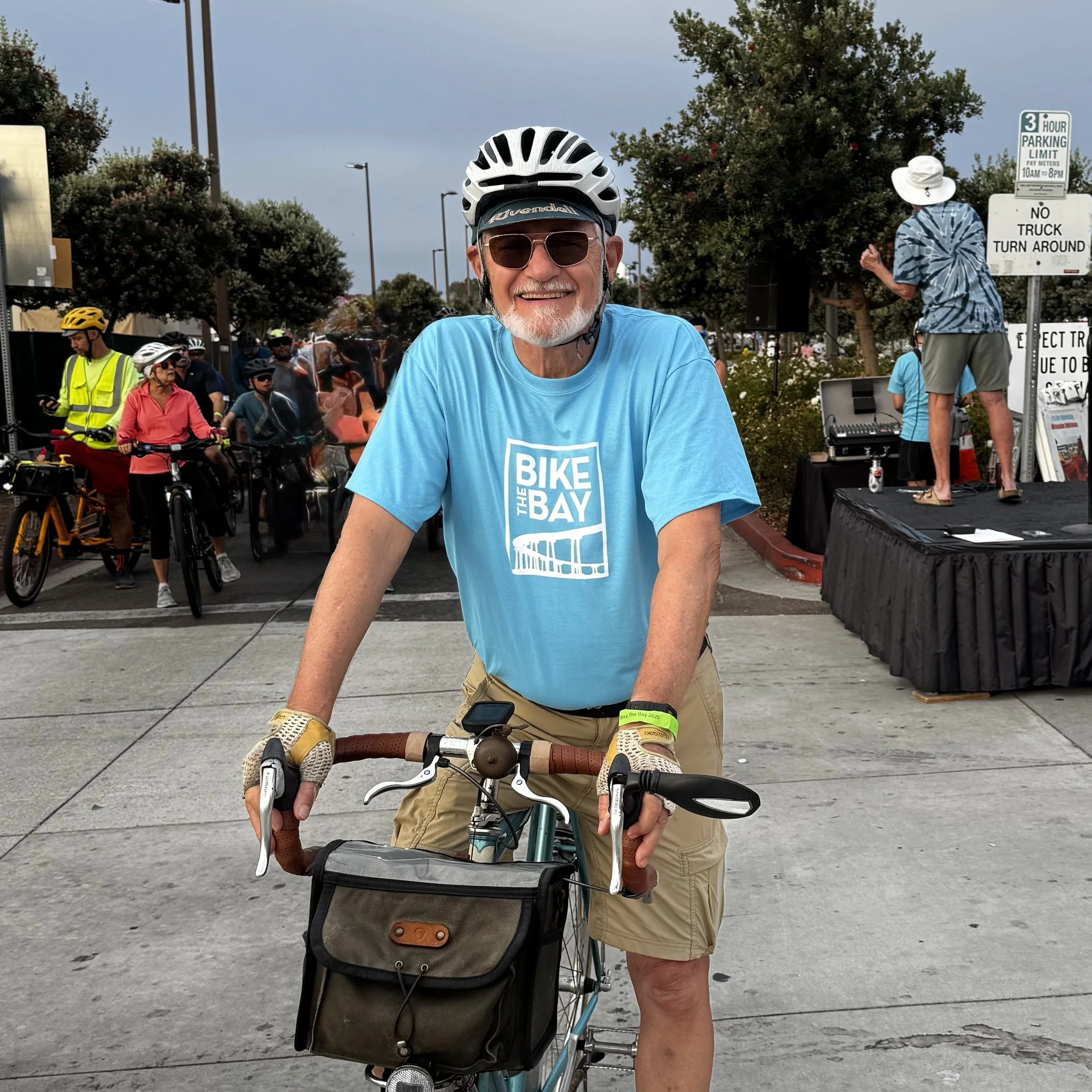 Older man wearing a blue T-shirt with 'Bike the Bay' logo, helmet, sunglasses, and gloves, standing with his bicycle at a cycling event, smiling at the camera, with other cyclists and event setup in the background.