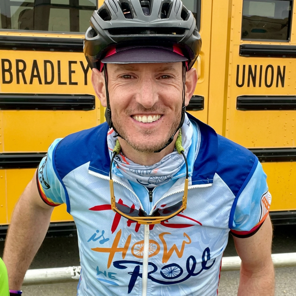 Smiling man wearing a blue cycling jersey and helmet, standing in front of a yellow school bus.