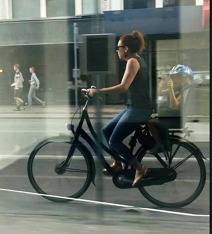 A woman riding a bicycle on a city street with a child seated in a rear child seat, taking a photo with a smartphone. Pedestrians walk in the background.