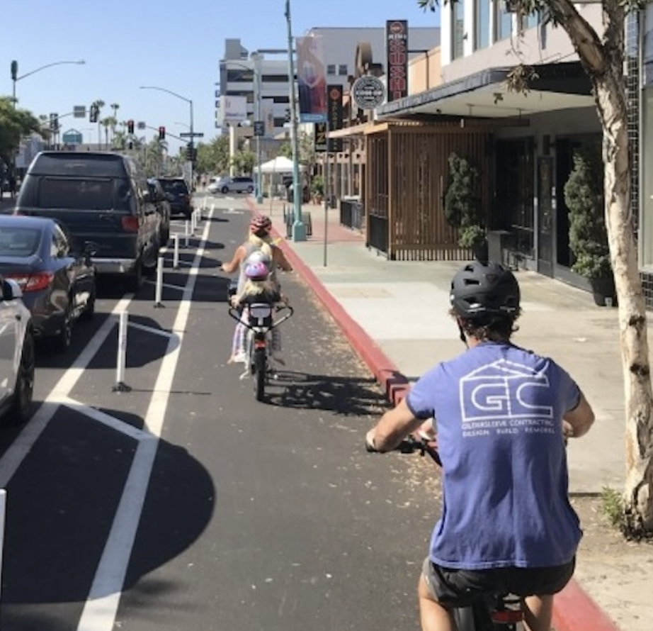 People riding bicycles on a city street with parked cars and storefronts.