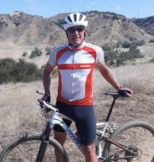 Man in cycling gear with helmet and sunglasses standing with a mountain bike in a dry, hilly landscape