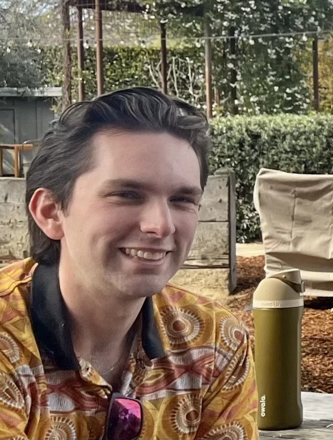 Man wearing a blue bicycle helmet and green T-shirt that says 'LIFE BY BIKE,' sitting on a bicycle on a city sidewalk with crosswalk and street in background.