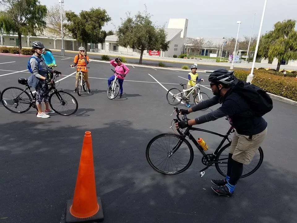 A group of people with bicycles gather in a parking lot during a cycling lesson, with one person holding a bike near an orange traffic cone.