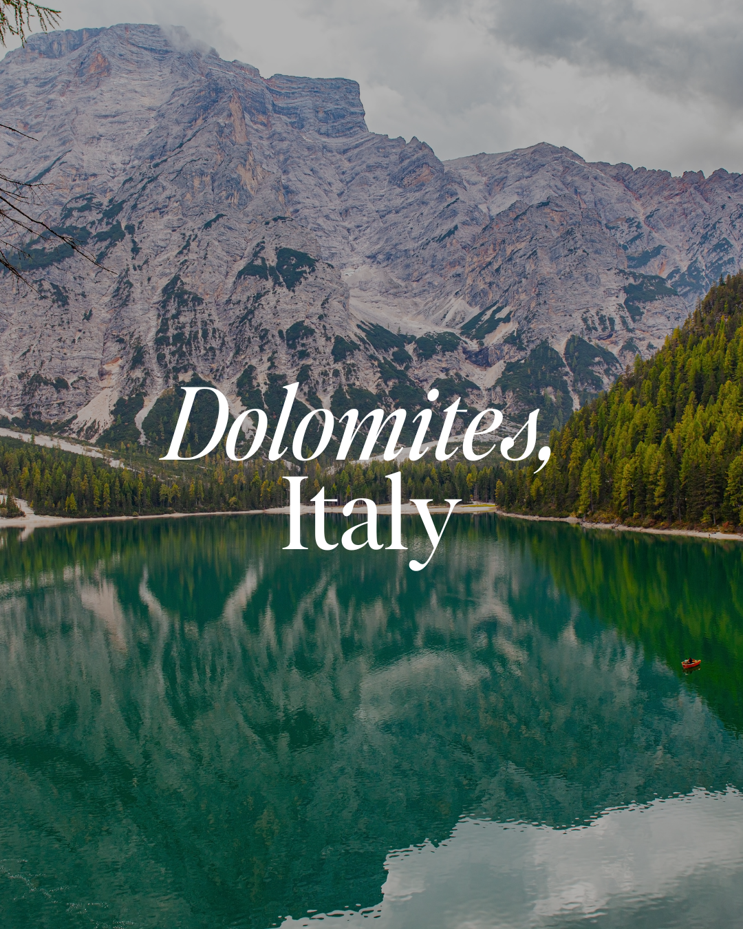 Scenic view of a lake surrounded by green forests with tall mountainous cliffs in the background. The water is calm and reflects the mountains and forest. Text overlay says 'Dolomites, Italy'.