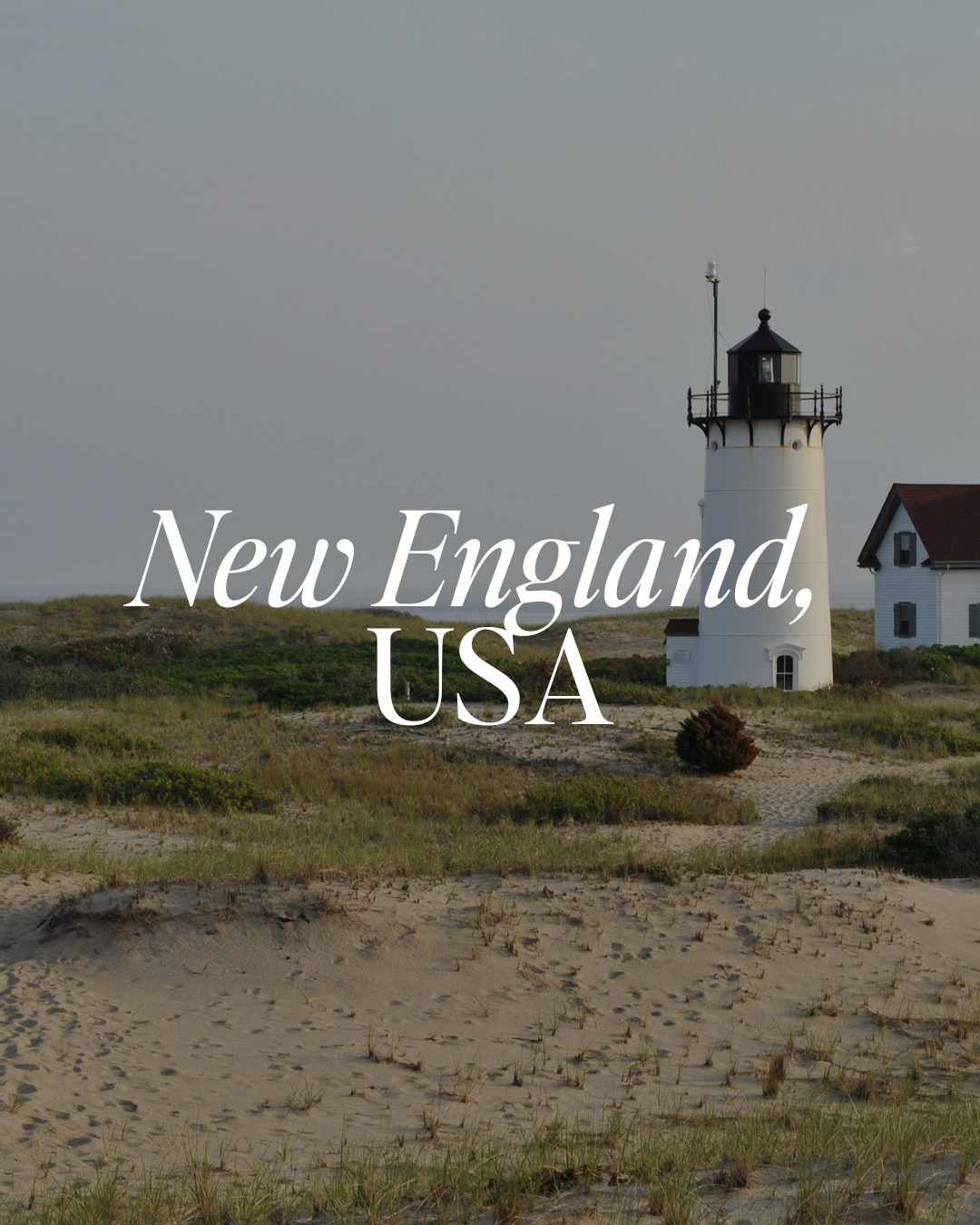Lighthouse on sandy shore with grassy dunes and a small house in the background, under cloudy sky. Text overlay says 'New England, USA'. 