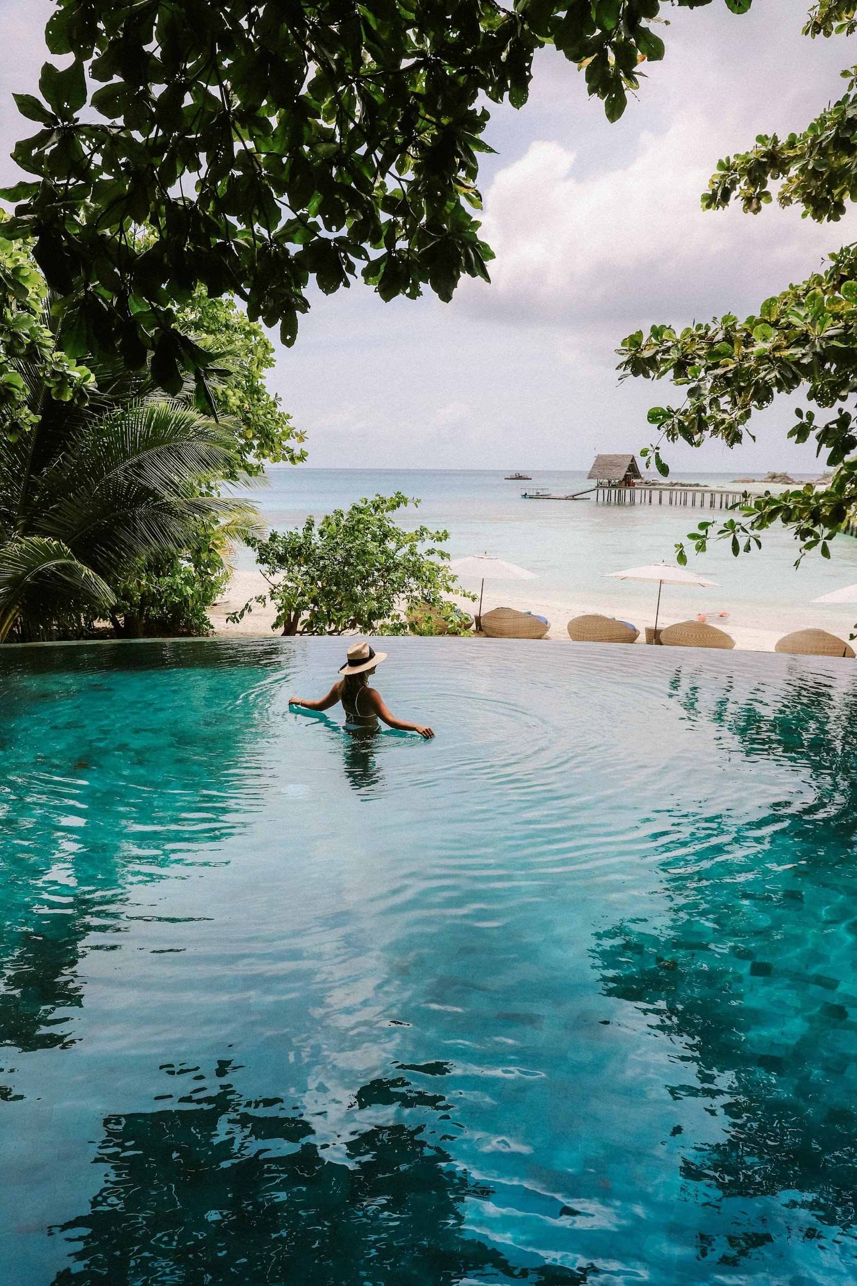 A woman in a swimsuit and straw hat stands in an infinity pool overlooking a beach with umbrellas, palm trees, and a pier in the distance.